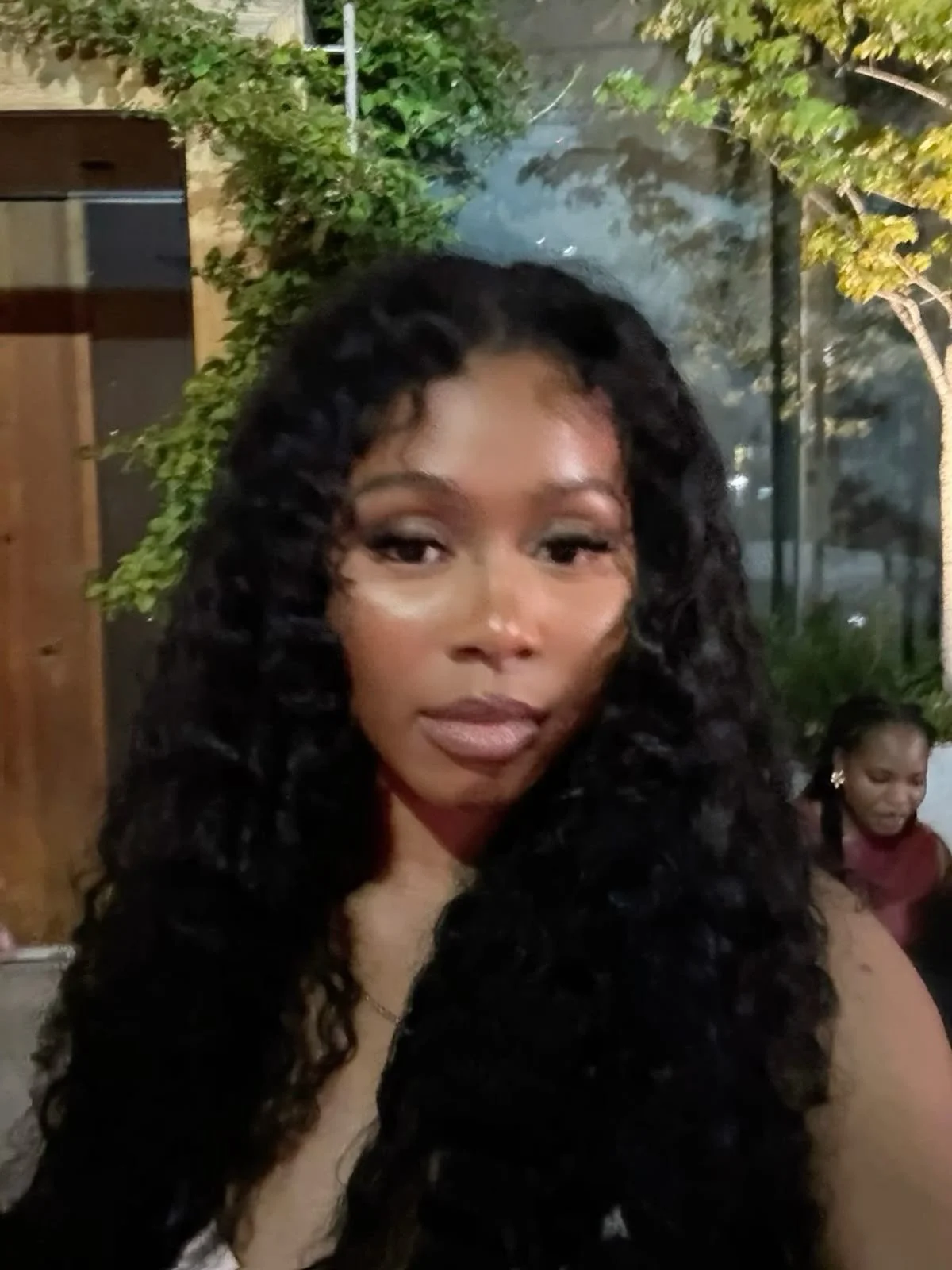 A woman with long, curly black hair, makeup, and a confident expression, standing outdoors at night near trees and a glass building.
