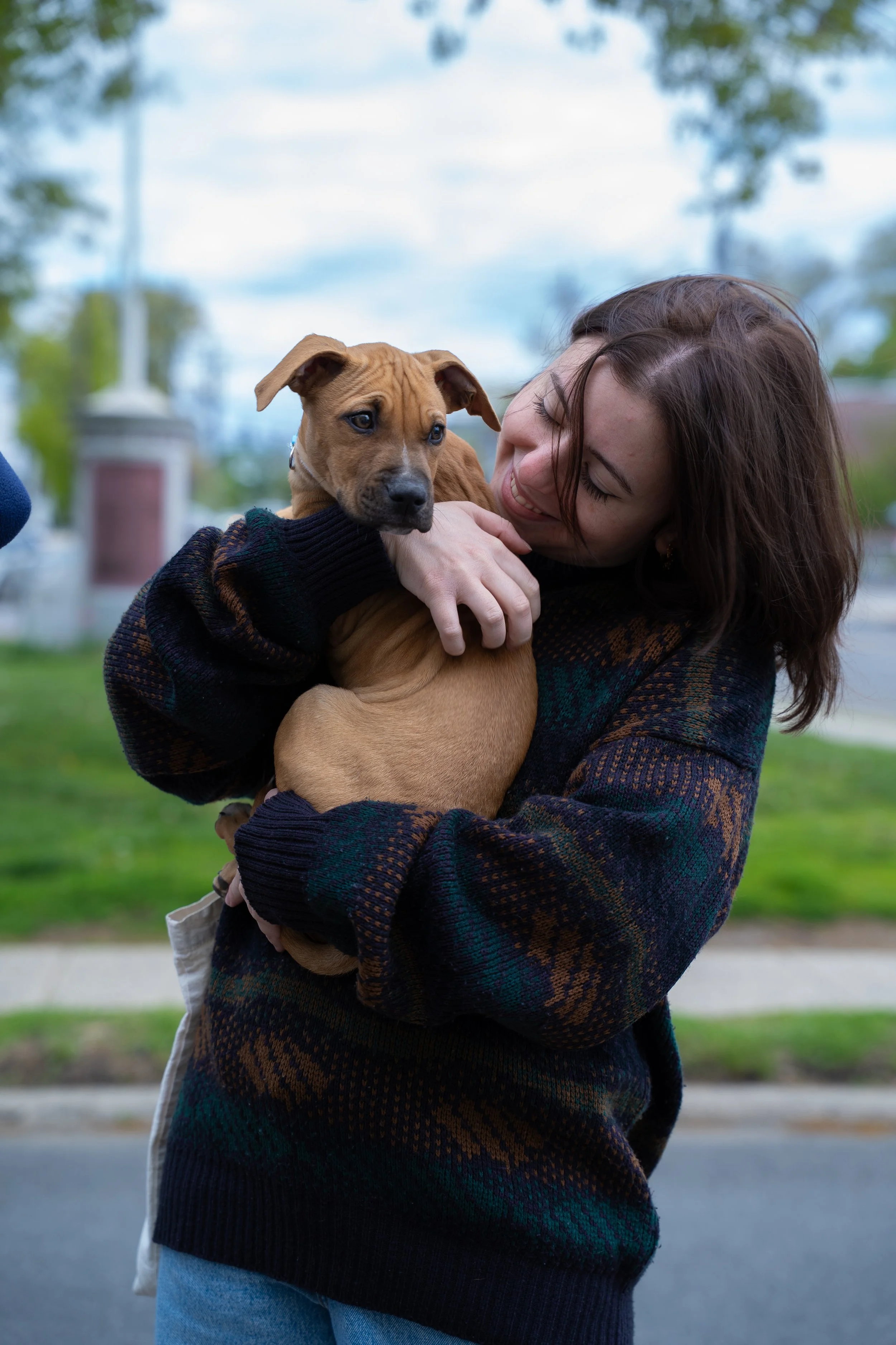 A woman with brown hair smiling as she holds a brown puppy at a dog adoption event