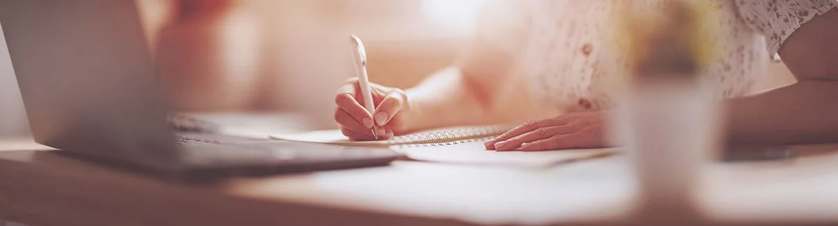 A person writing in a notebook on a wooden table next to a laptop and a blurred potted plant.