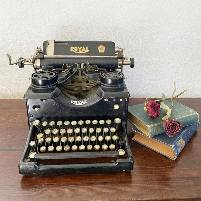 Vintage black Royal typewriter with white keys on a wooden table, next to three stacked books with a dried red rose on top.