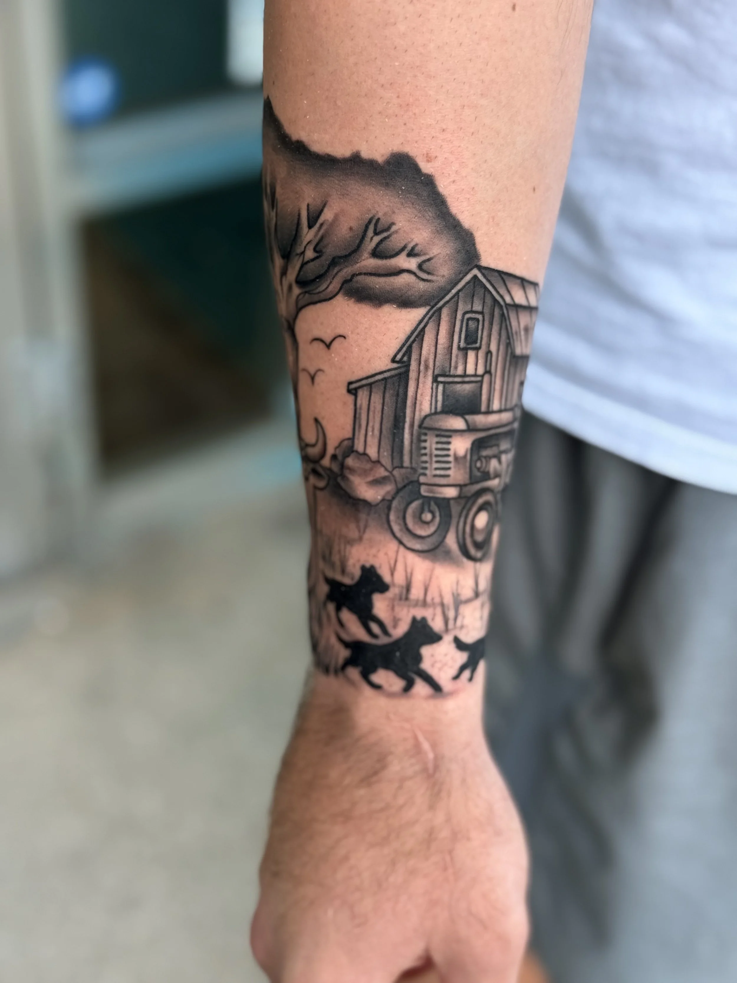 Tattoo of a rural farm scene on an arm, featuring a barn, a tractor, a wolf pack, flying birds, a leafless tree, and clouds.