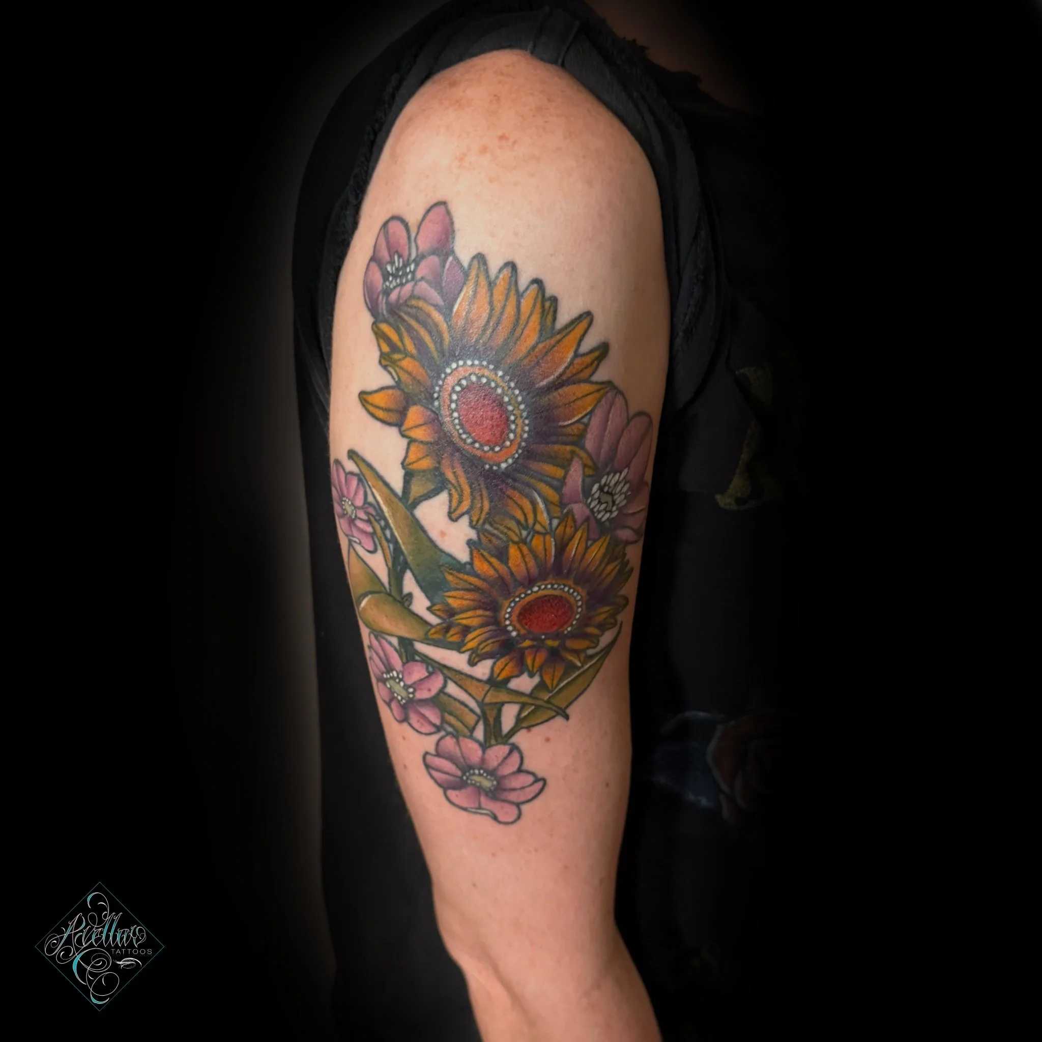 Colorful floral tattoo featuring sunflowers, pink flowers, and green leaves on person's upper arm.