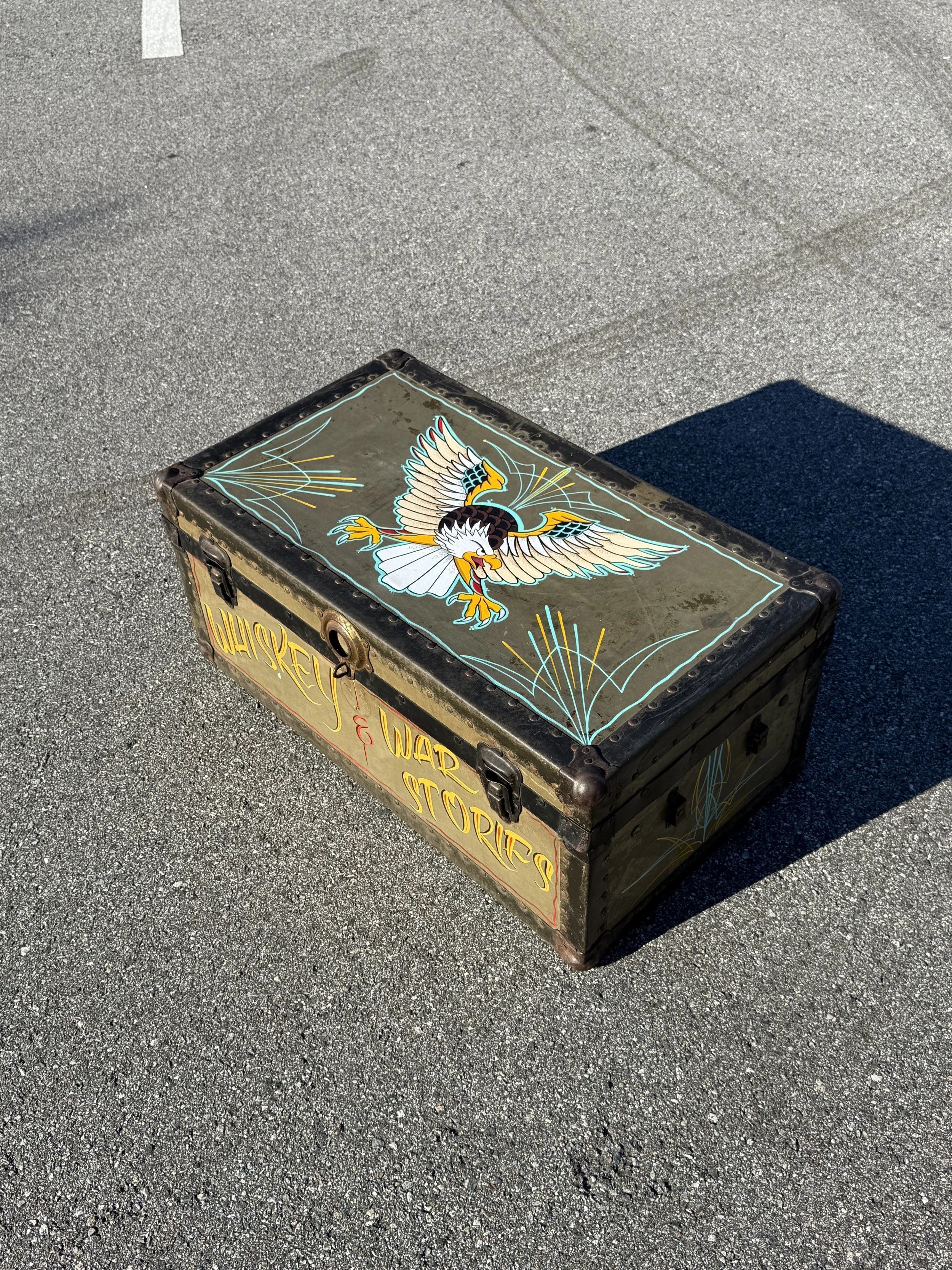 A vintage WW2 foot locker painted with an American eagle in flight, surrounded by decorative lines, with the words "WISKEY & WAR STORIES" written on the side.