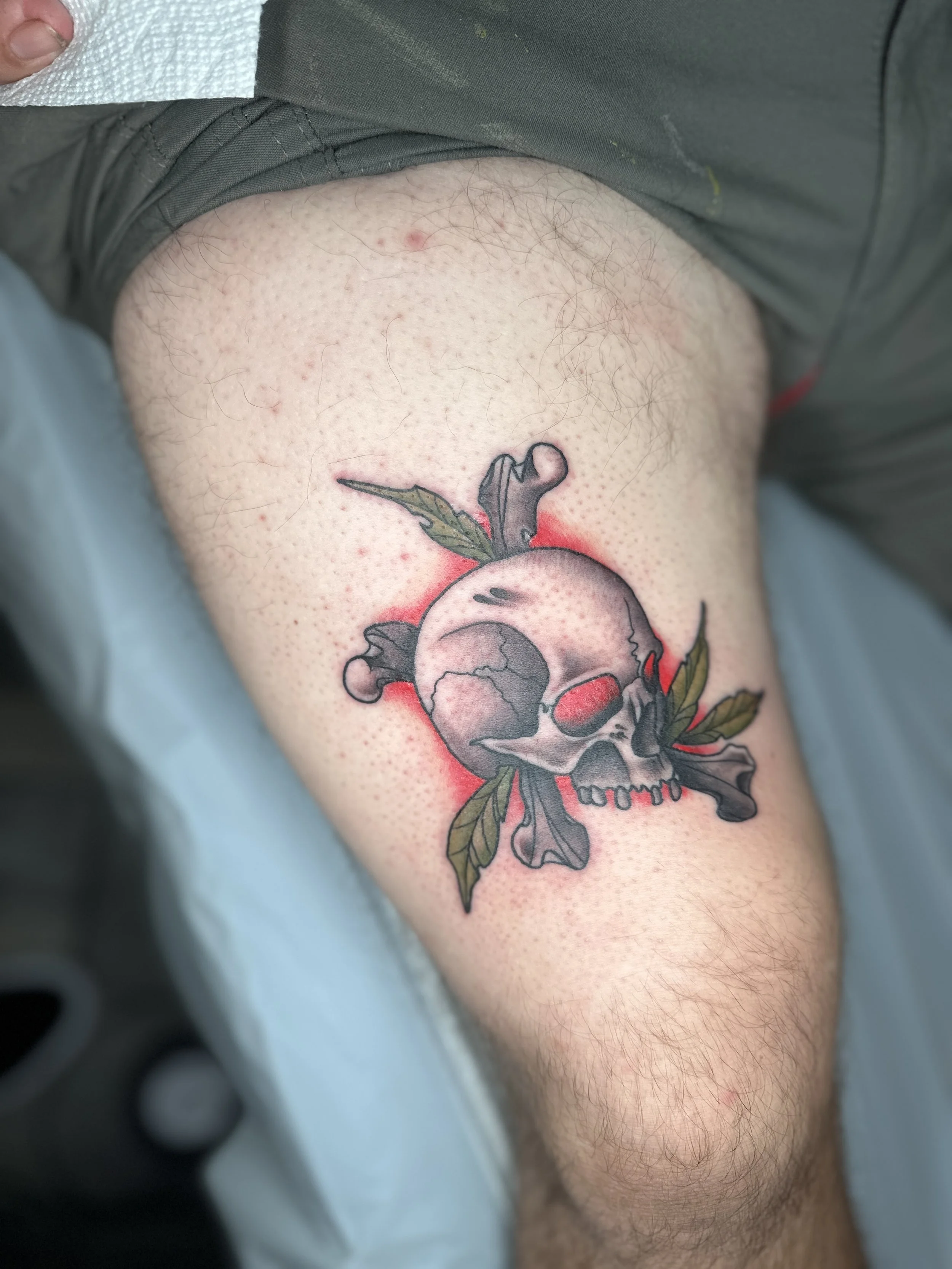 A fresh tattoo of a skull with a pinkish-red hue, surrounded by green leaves and black smoke, on a person's thigh.