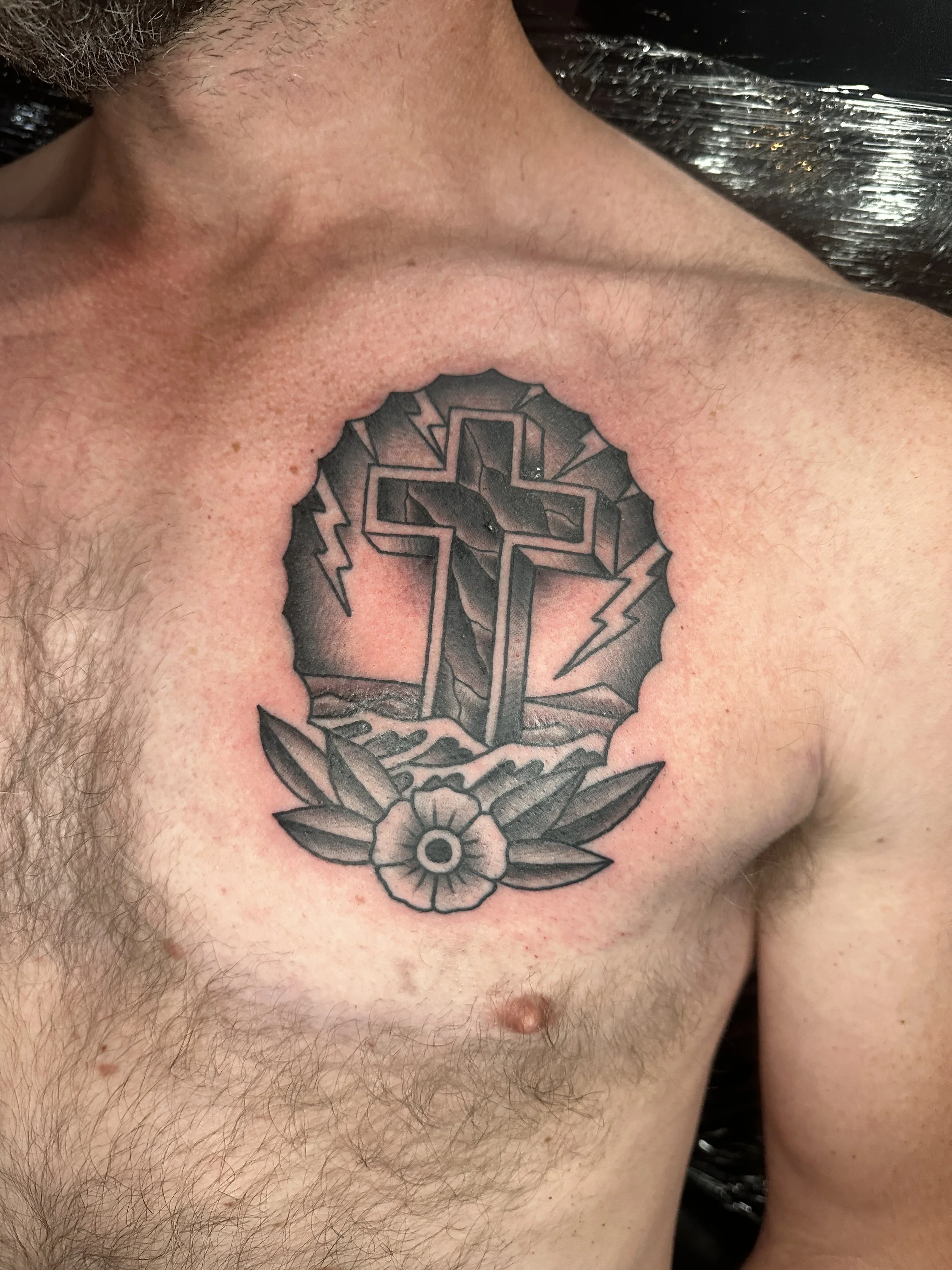 Tattoo of a cross with lightning bolts behind it, surrounded by a circular border with a flower at the bottom and leaves.