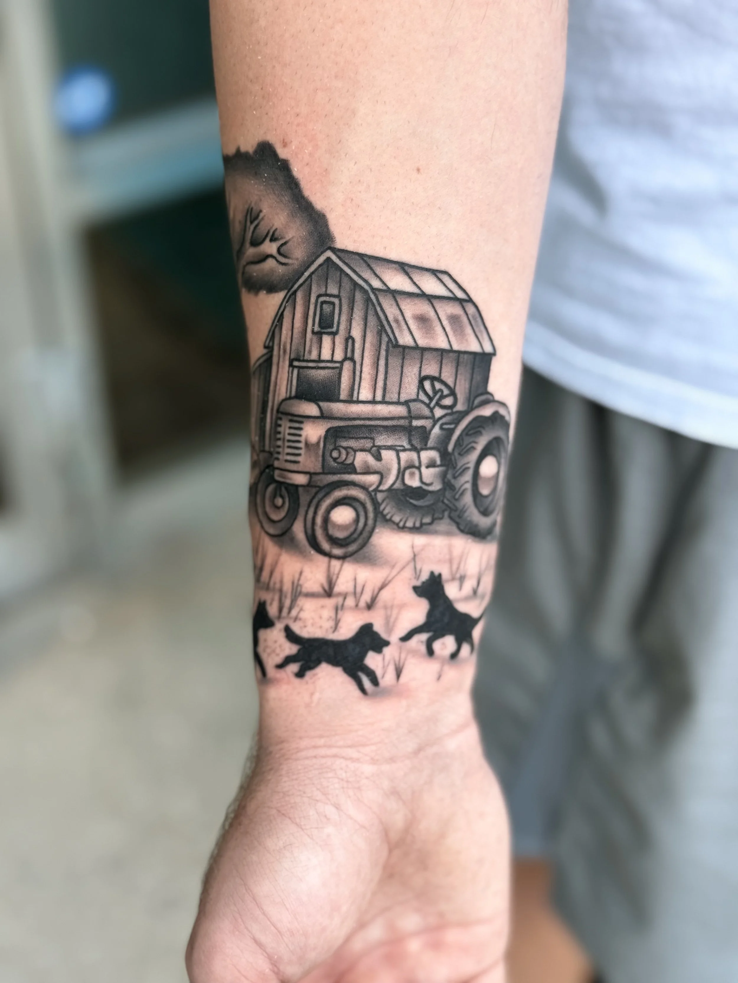 Tattoo of a barn with a tractor in front and silhouettes of two dogs running in front, with a tree and cloud in the background.