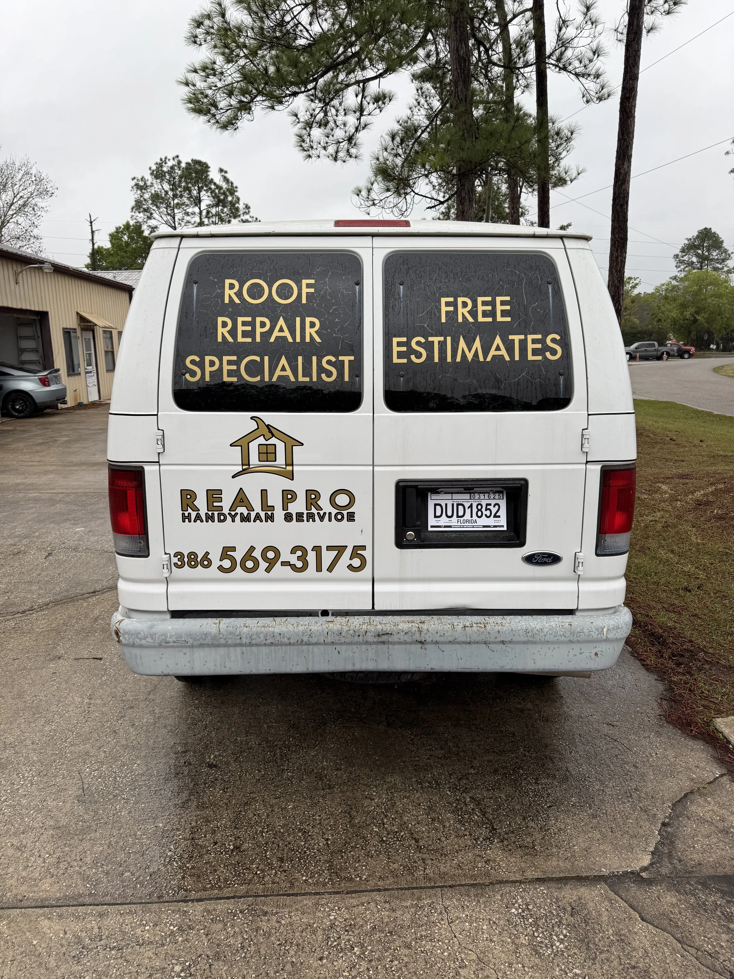 Rear view of a white handyman service van advertising roof repair with large windows displaying the text 'Roof Repair Specialist' and 'Free Estimates'. The company logo, 'Realpho Handyman Service', phone number, and a Florida license plate are also v