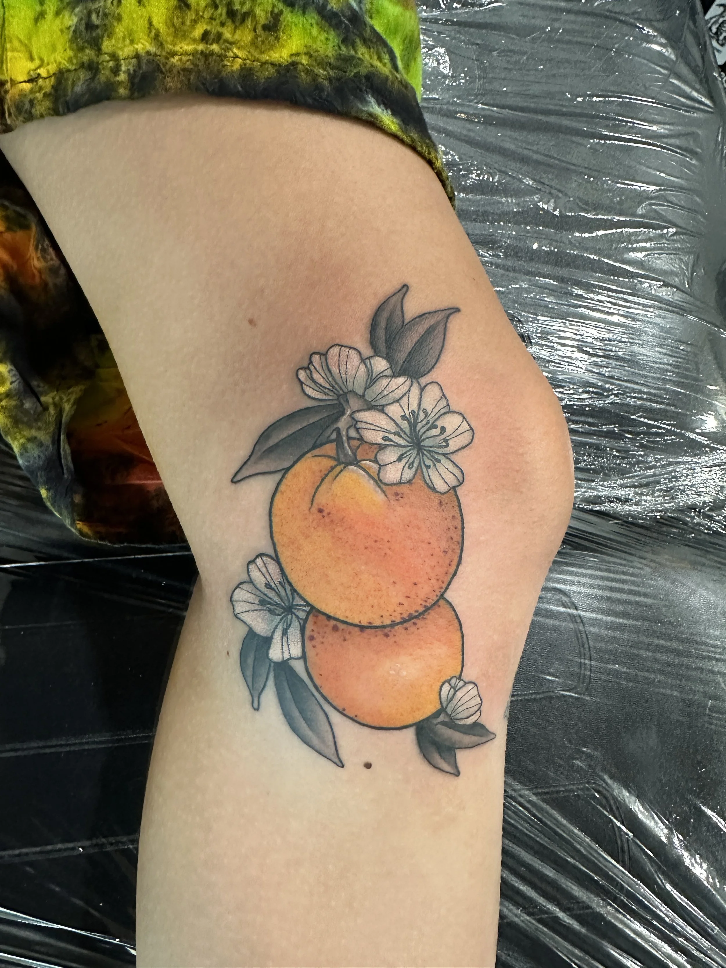 Tattoo of two oranges with leaves and blooming flowers on a person's thigh.