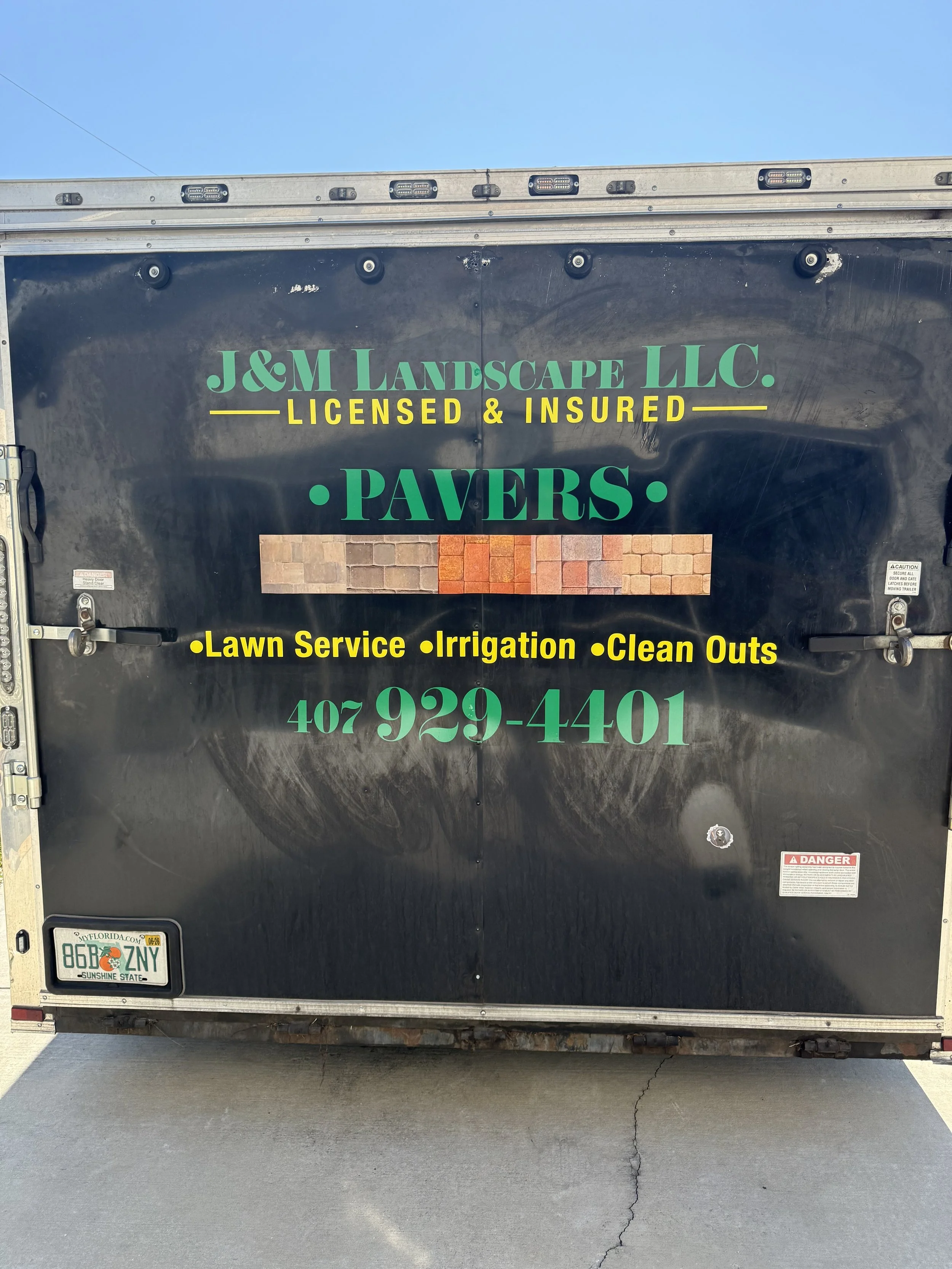 Black trailer with colorful text advertising J&M Landscape LLC, licensed and insured. Services include pavers, lawn service, irrigation, and clean outs. Phone number 407-929-4401. Small brick landscape design in the center of the trailer.