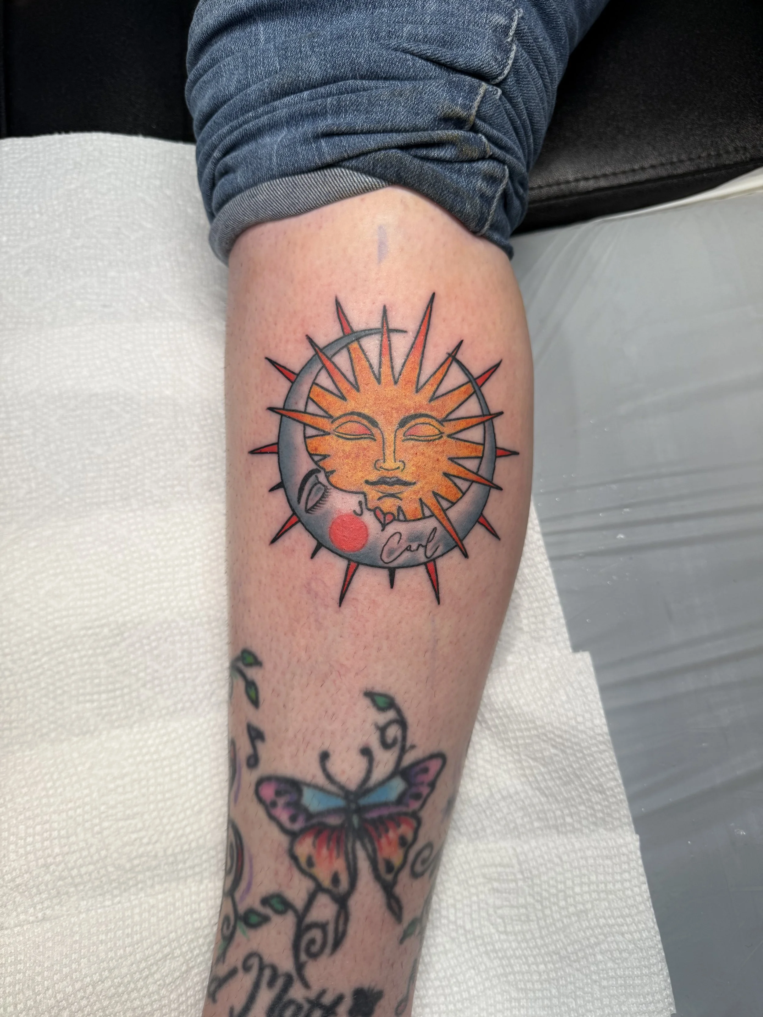 Tattoo of the sun with a human face, surrounded by a crescent moon with a human face and small red circles, on a person's upper leg.