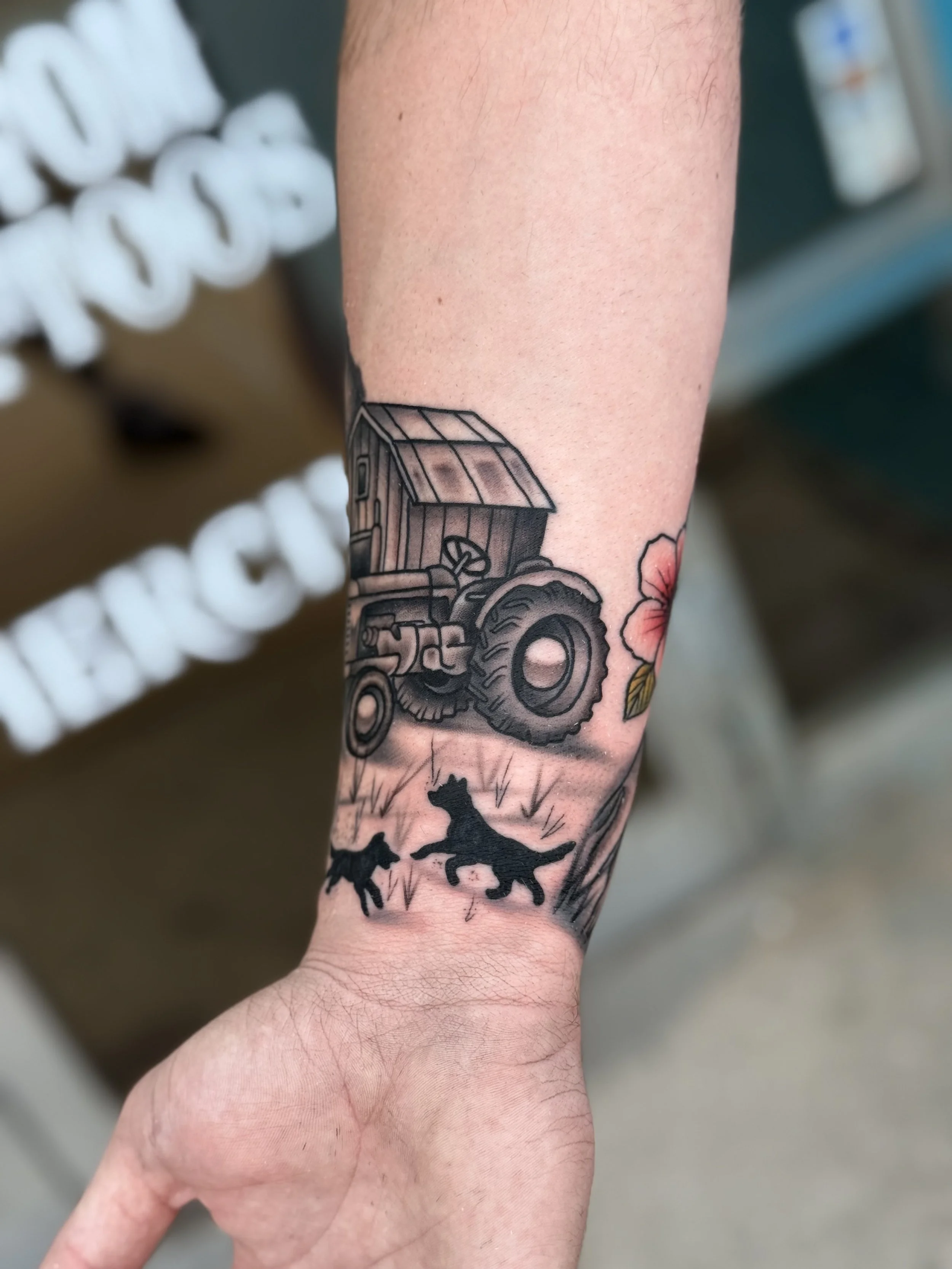 Tattoo on a person's forearm depicting a tractor, a pink flower, and silhouettes of three dogs running on grass.