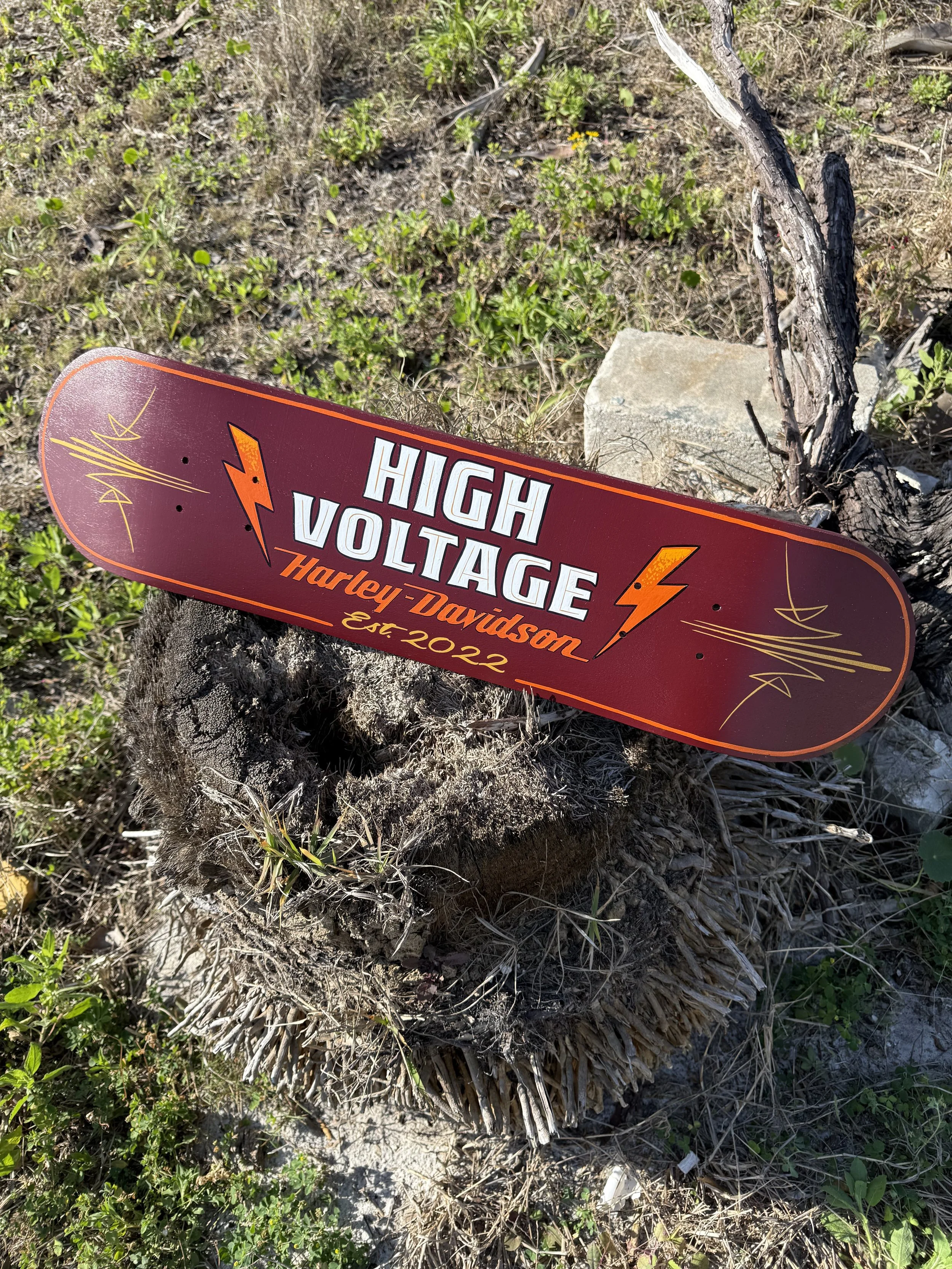 A custom painted skateboard deck that reads "High Voltage Harley-Davidson Est. 2022" with lightning bolt graphics, placed on a tree stump outdoors.
