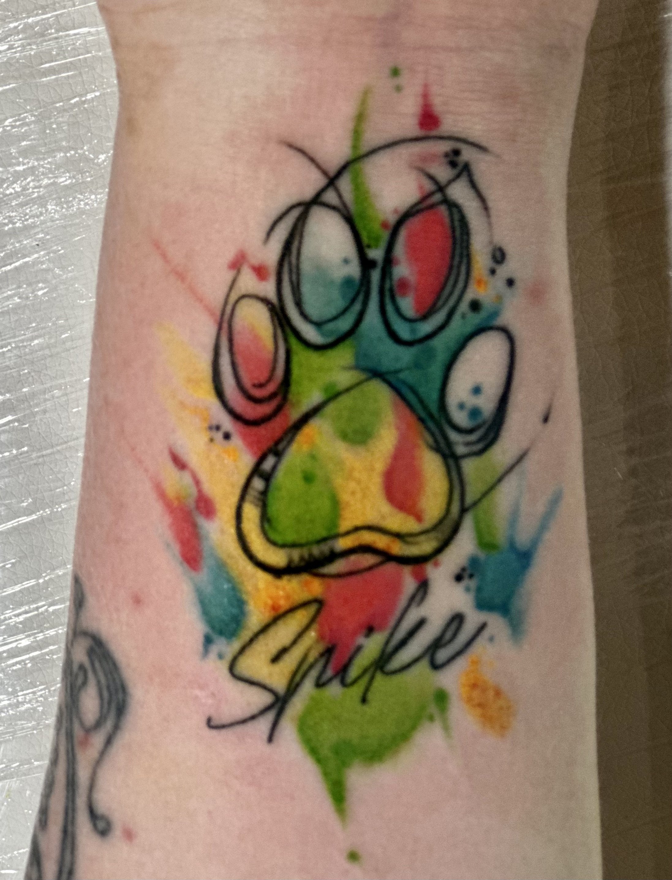 Tattoo of a dog’s paw with colorful watercolor splashes and the word 'Smile' written below.