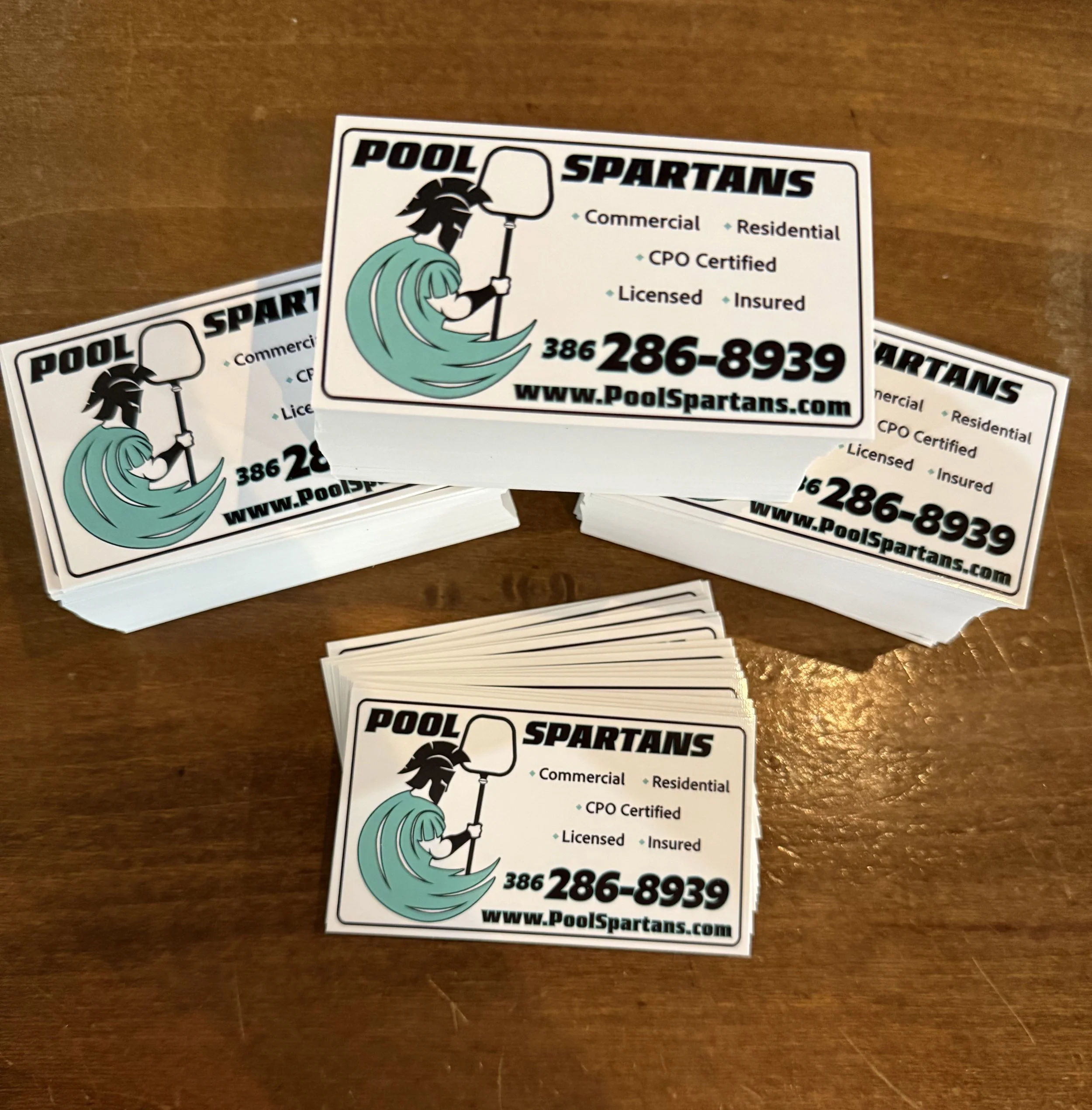 Stacks of business cards for Pool Spartans on a wooden surface. The cards feature a logo of a Spartan head with a pool net, and text displaying contact information and services offered, including phone number 386-286-8939 and website www.PoolSpartans