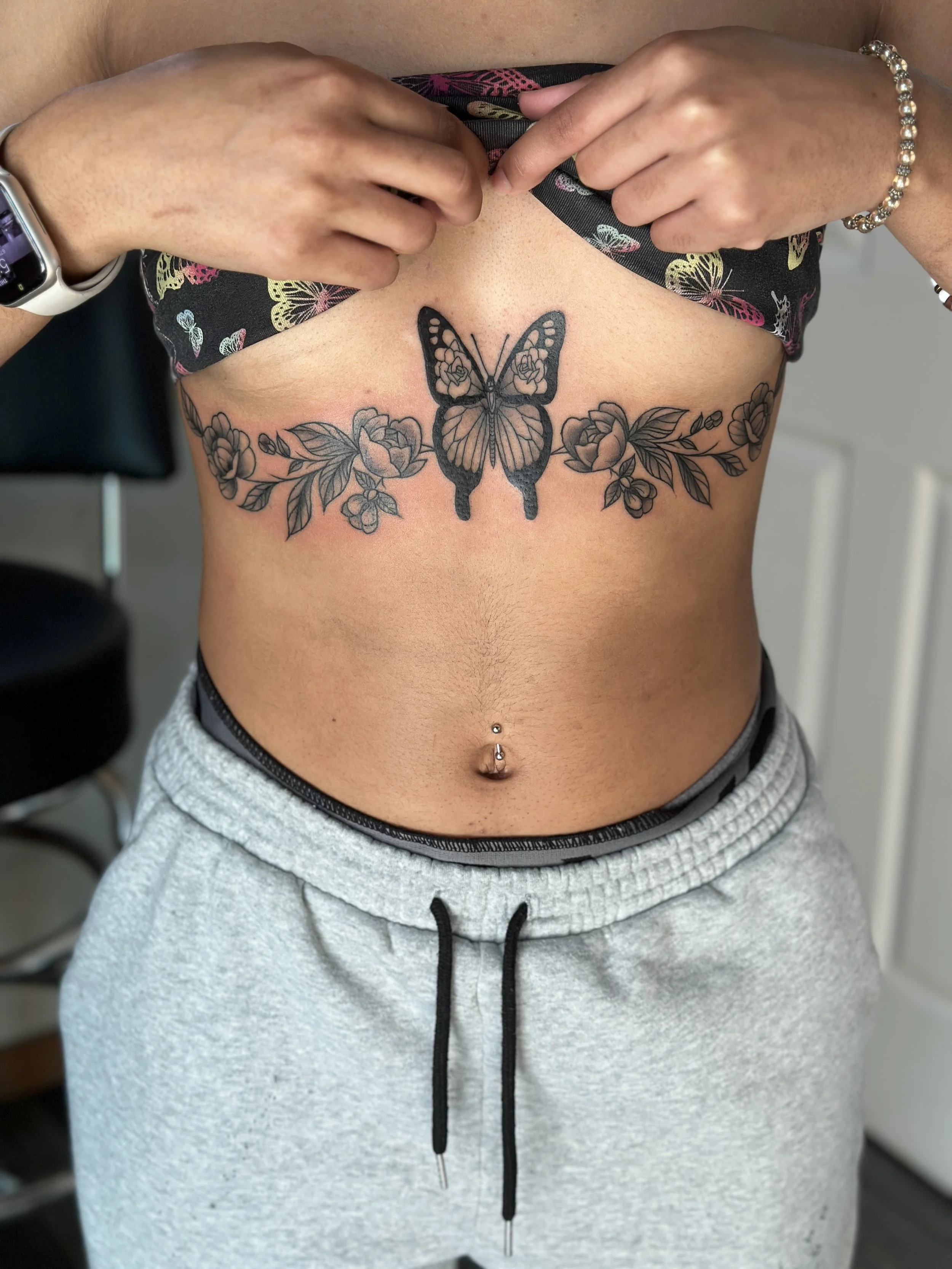 A person lifting their shirt to show a tattoo of a butterfly with flowers on their chest.