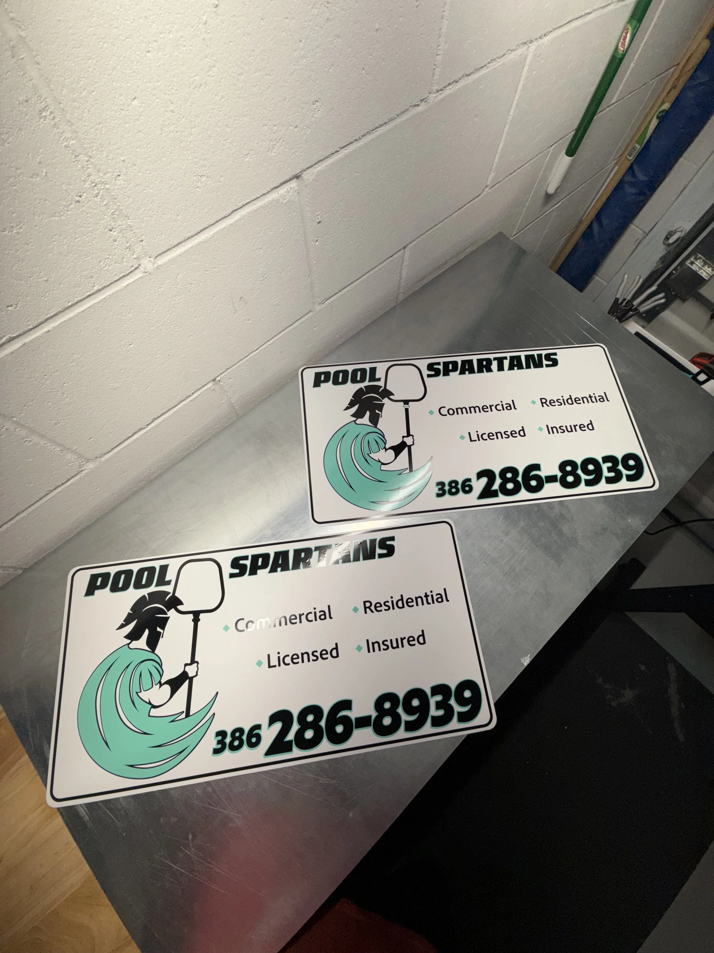 Two identical pool service magnetic signs signs on a metal table, showing a cartoon figure of a person with a towel, holding a pool net, with text including the company name "Pool Spartans," services offered, and a phone number.