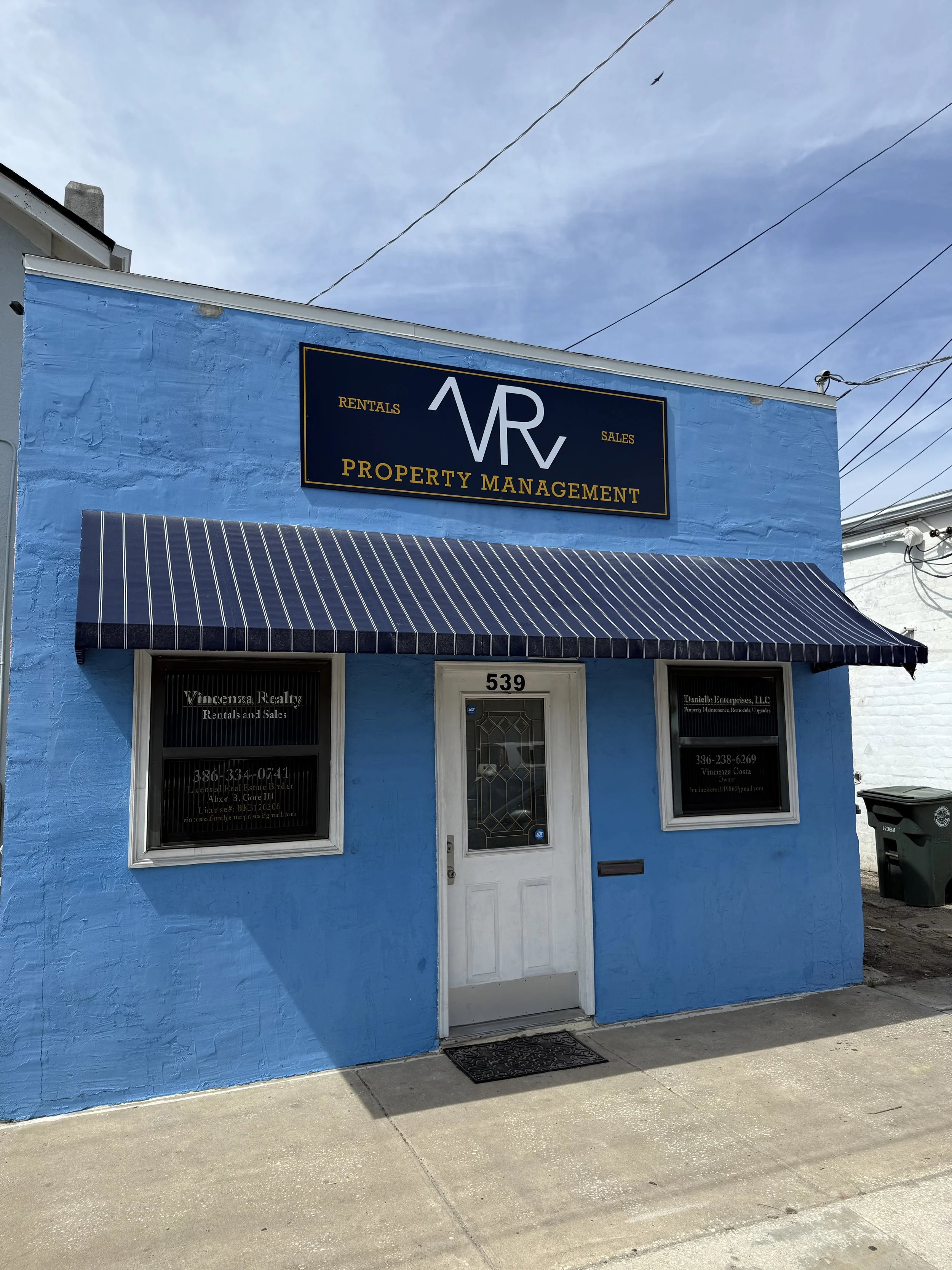 Blue building with a sign that reads 'AVR Property Management' and includes words 'Rentals' and 'Sales'. There is an awning over the entrance, which is a white door with glass, and two windows on either side. The left window has a sign for 'Vincenza