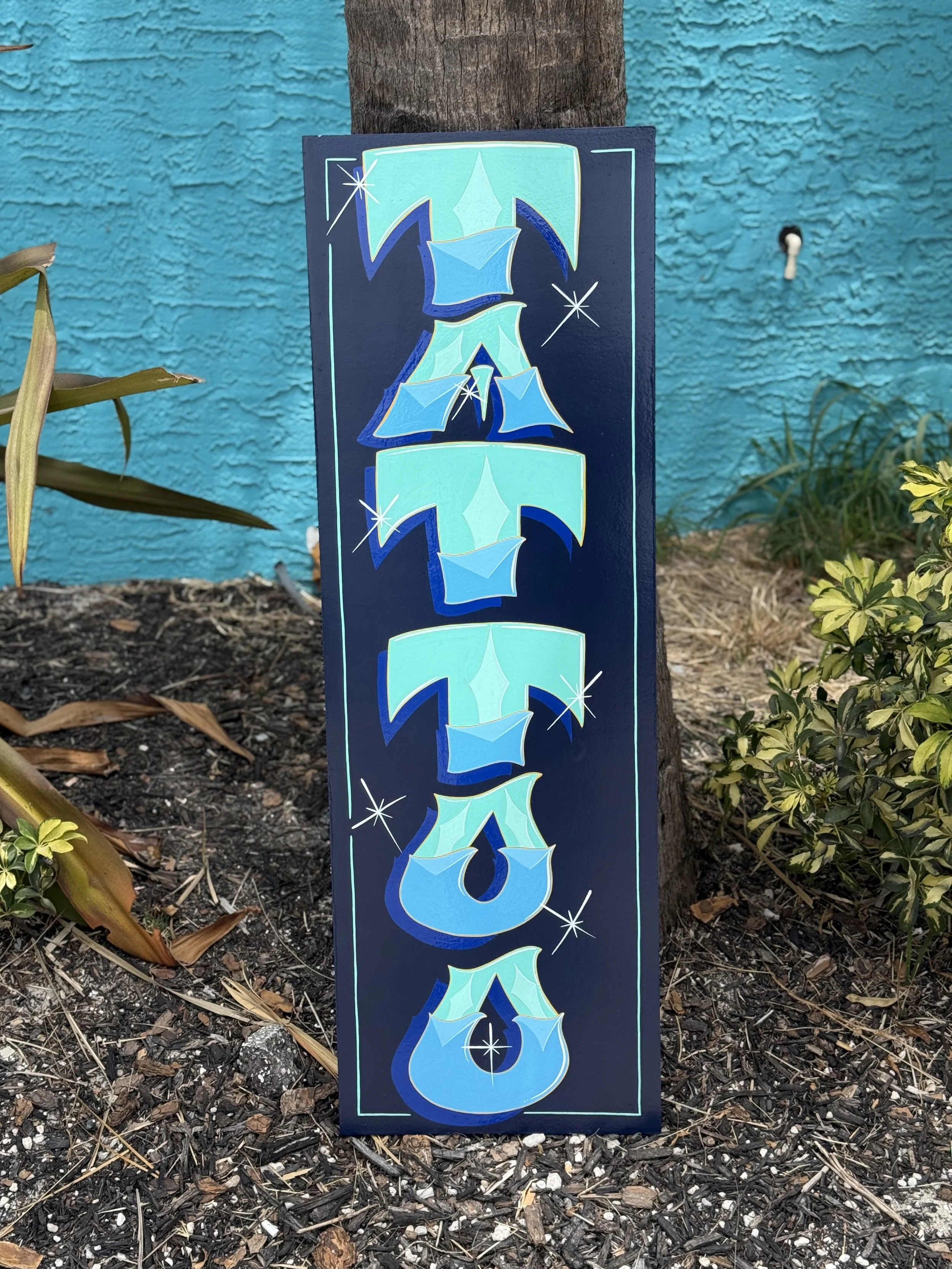 Colorful vertical sign with stylized blue and turquoise letters spelling 'VISA', decorated with stars, attached to a tree outside near plants and soil, with a textured turquoise wall in the background.