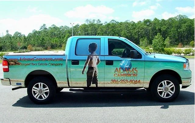 Blue pickup truck with a boy's silhouette full wrap graphic on the side and advertising for Adams Cameron real estate company.