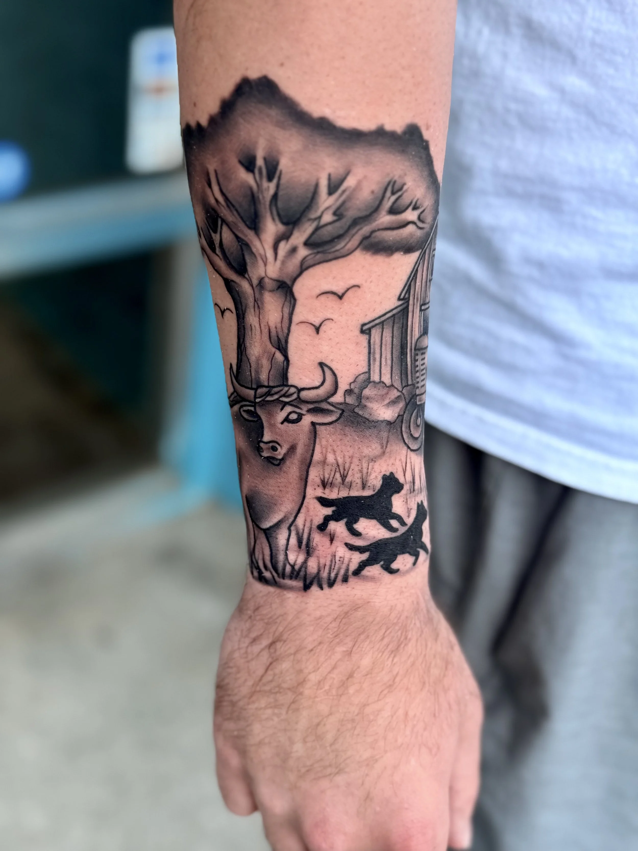 Tattoo of a deer head with large antlers, a tree, a barn, and two small people or animals walking near the grass.