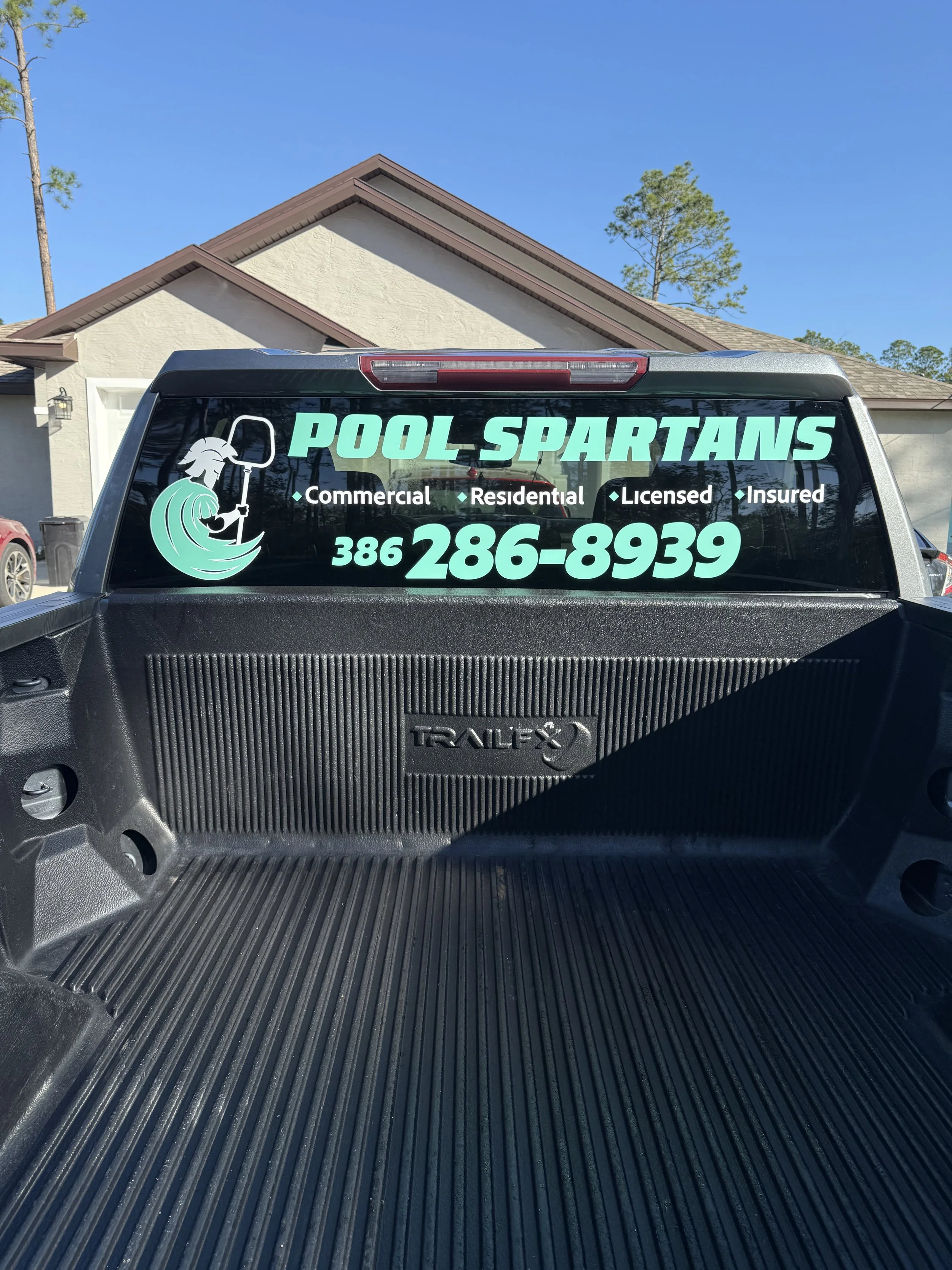 Back window of a pickup truck with a "Pool Spartans" advertisement, featuring a logo of a warrior with a shield and spear, and contact information in bright green text.