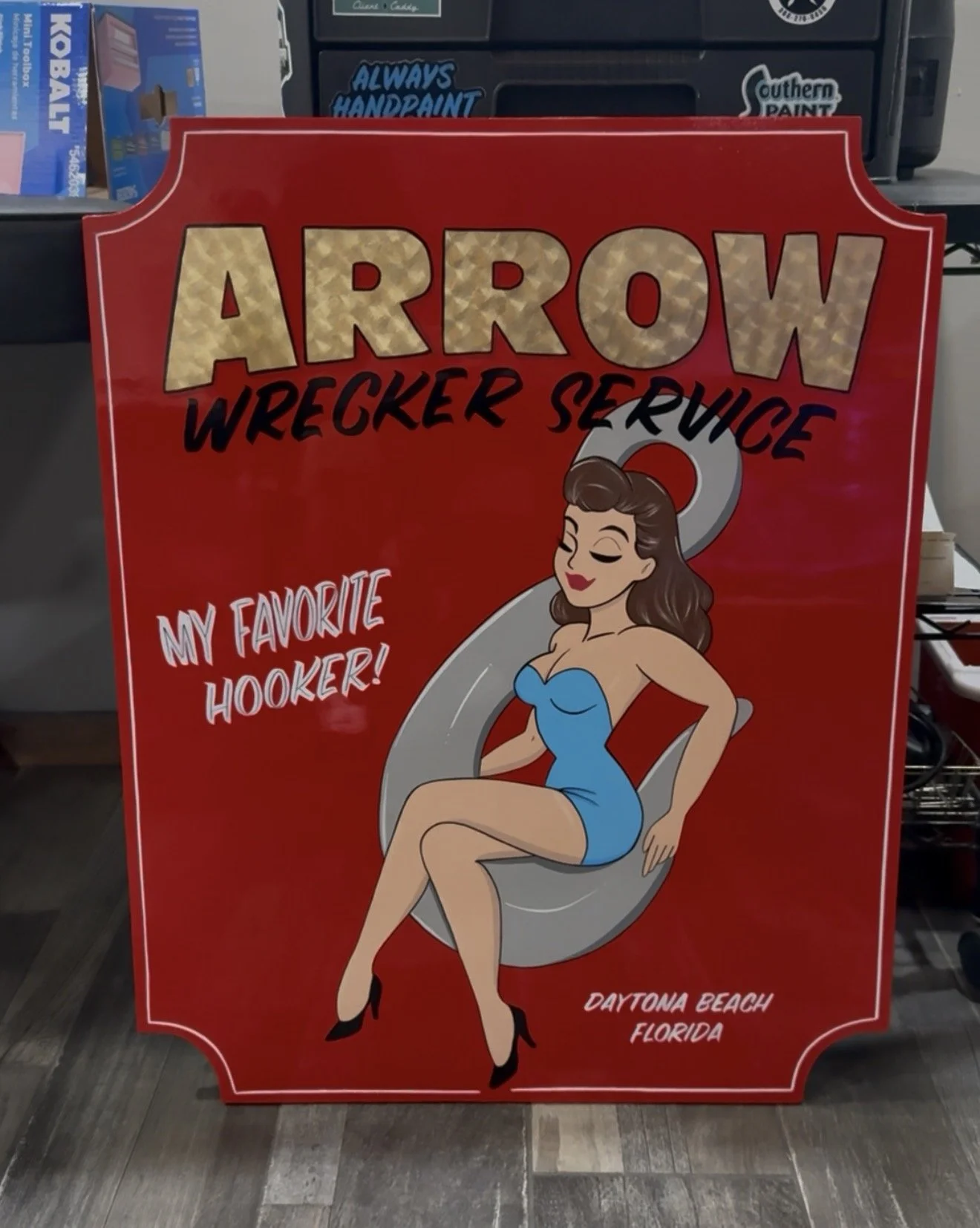 Red sign with cartoon woman in blue dress sitting on a boat prop, with text 'Arrow Wrecker Service,' in gold leaf  'My Favorite Hooker!,' and 'Daytona Beach, Florida.'