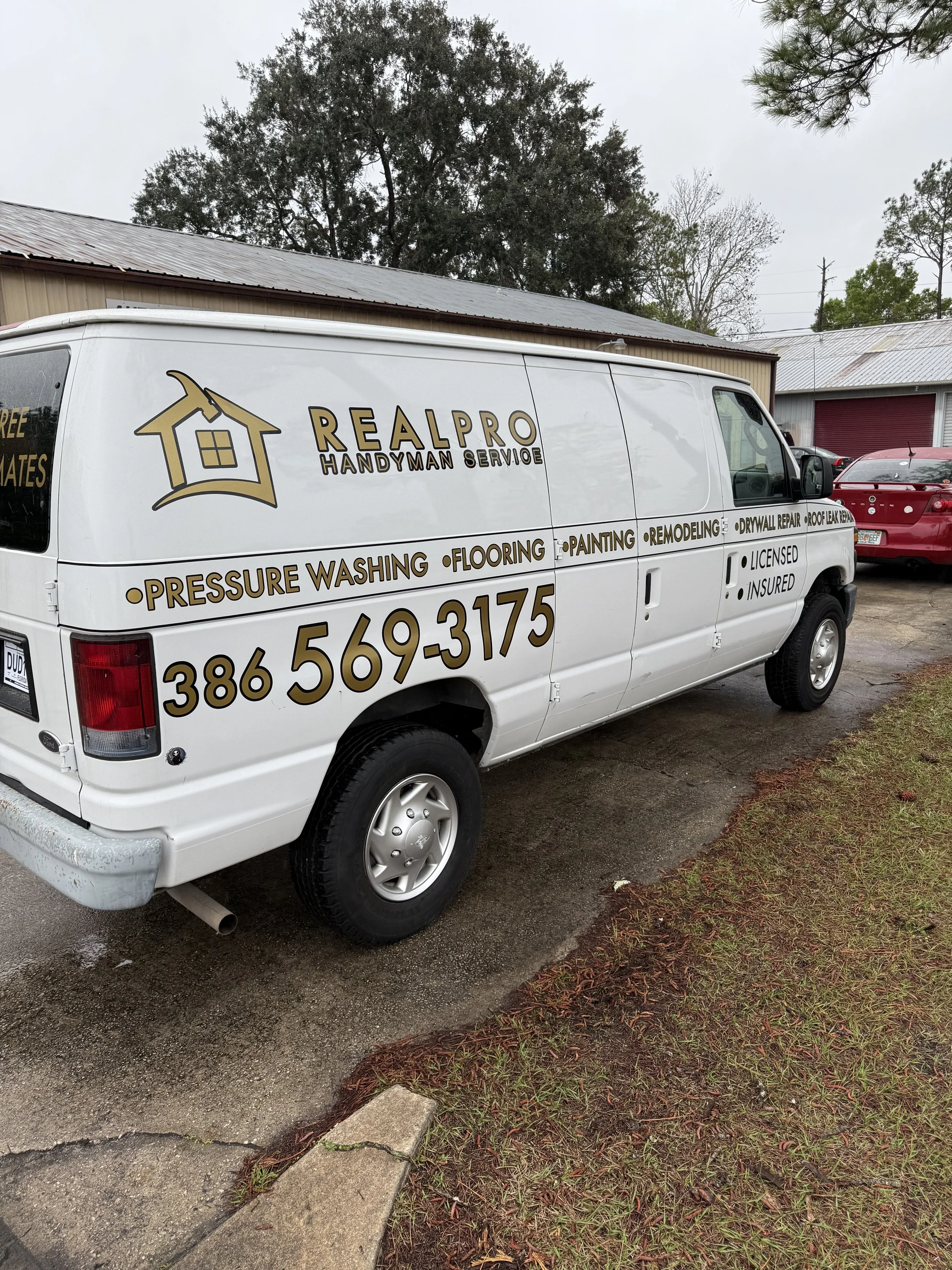 White van with business advertisement for Real Pro Handyman Service, including phone number, list of services like pressure washing, flooring, painting, remodeling, and repair services, parked outdoors near a building and other vehicles.