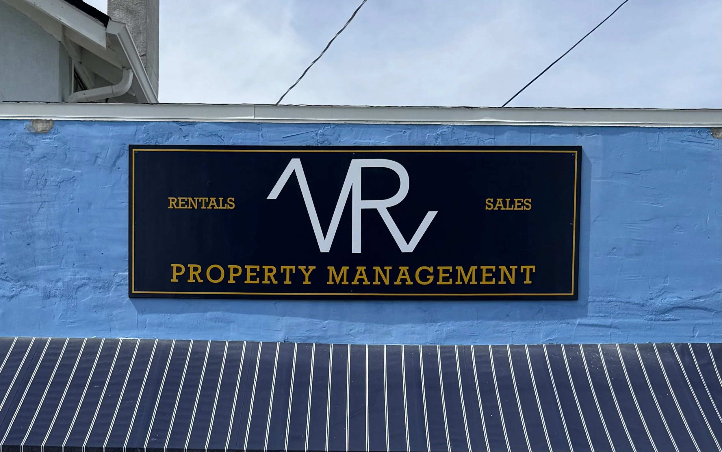 Sign for VR Property Management on a blue wall, with services in rentals, sales, and property management, and a striped awning below.