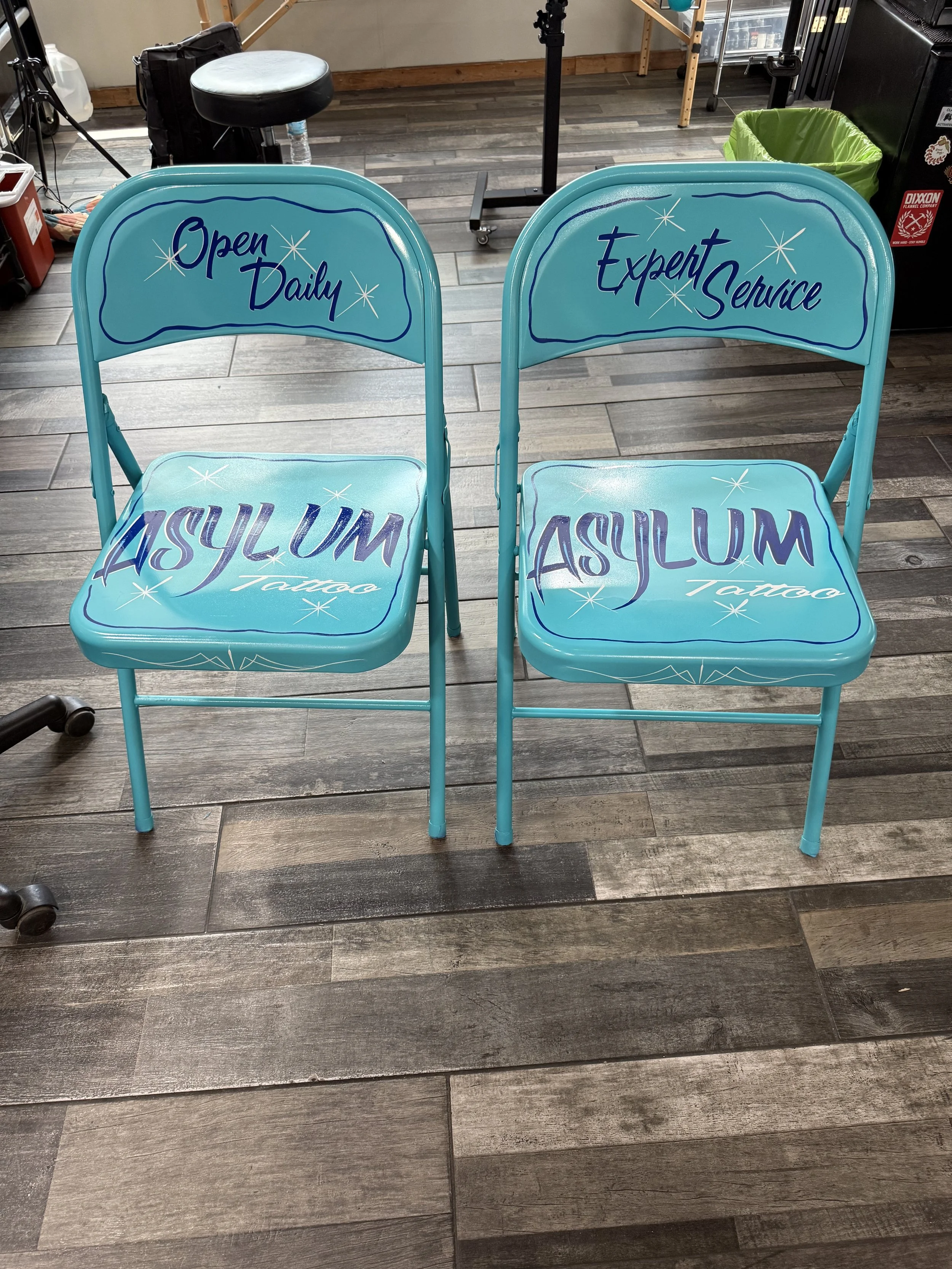 Two turquoise chairs with the words "Open Daily" and "Expert Service" on the backrests and "ASULUM TATTOO" written on the seats, placed on a wooden floor in a tattoo studio.