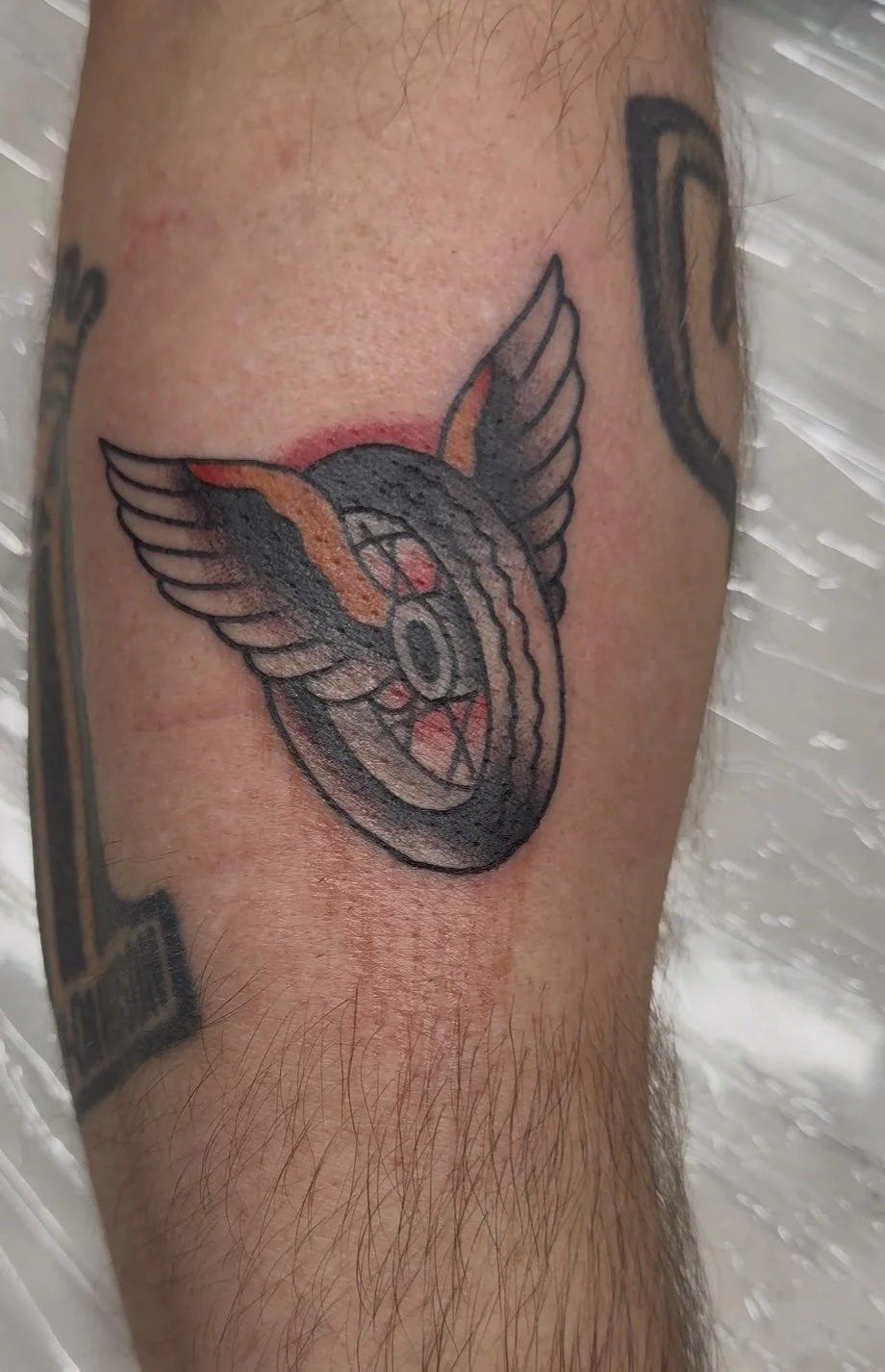 Tattoo of a stylized wolf's head with wings, featuring bold black outlines, red and orange accents, and a circular design in the center.