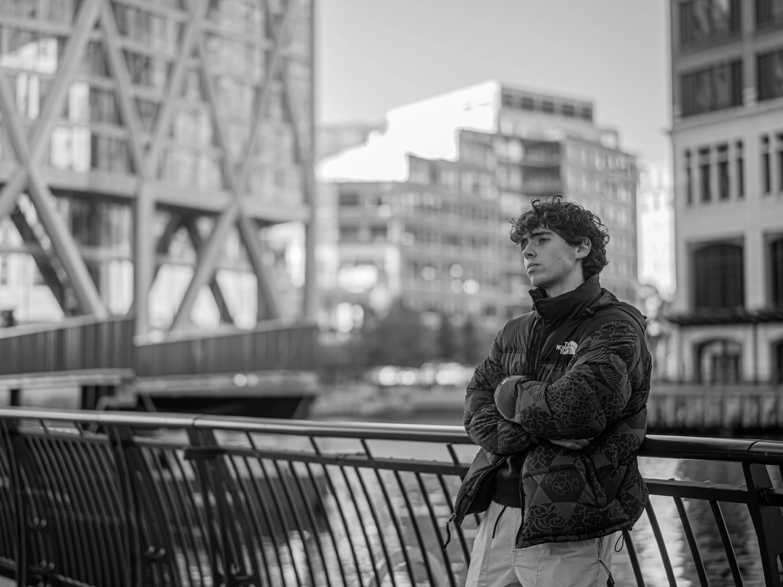 A young man with curly hair and a black North Face jacket, standing with arms crossed, on a bridge in an urban setting, with modern buildings in the background, in black and white.