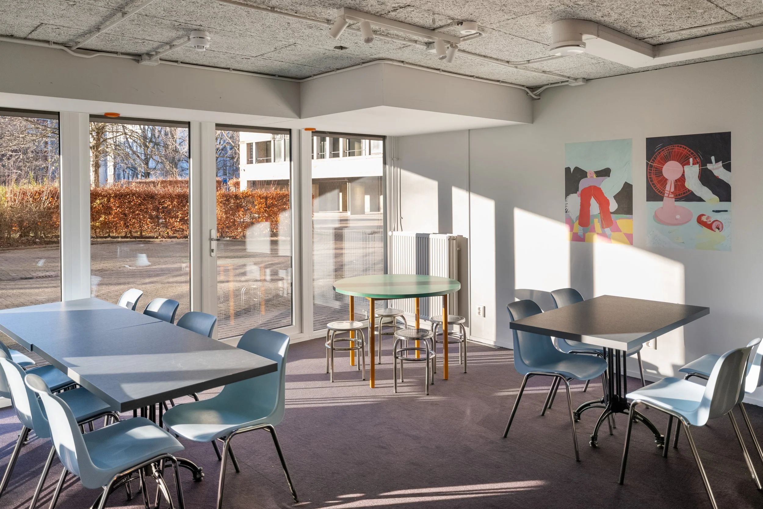 Empty room with large glass windows, modern tables and chairs, colorful abstract artwork on the wall, and sunlight streaming in.