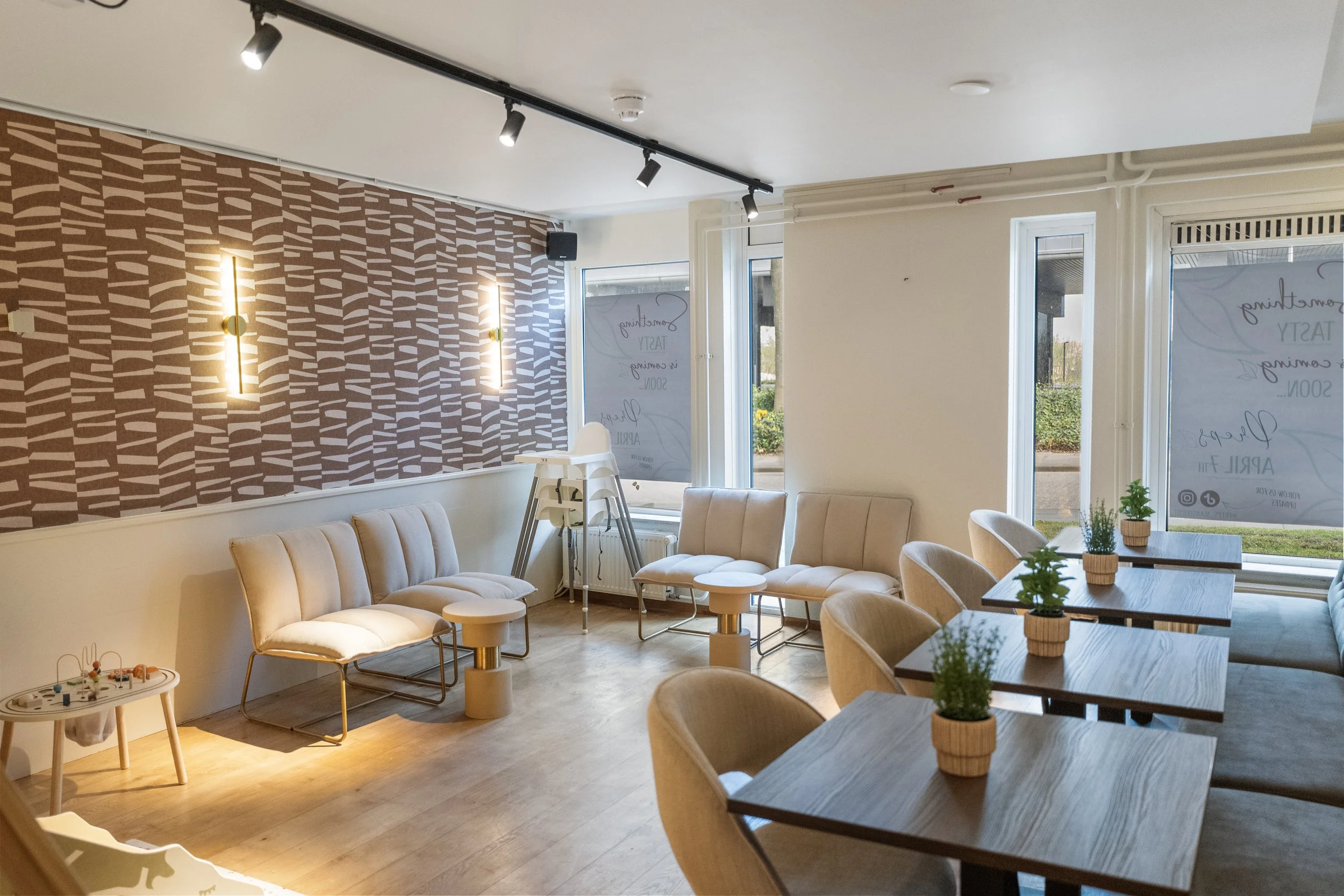 Interior of a modern cafe with beige chairs and small potted plants on wooden tables, large windows, and decorative lighting on a patterned accent wall.