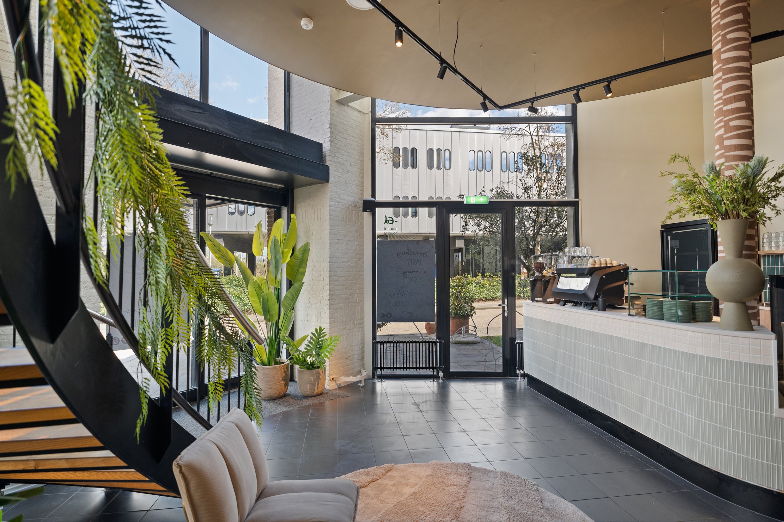 Interior of a modern coffee shop with large windows, black tile flooring, and a white tiled counter with a coffee machine, stacks of cups, and decorative plants in vases.