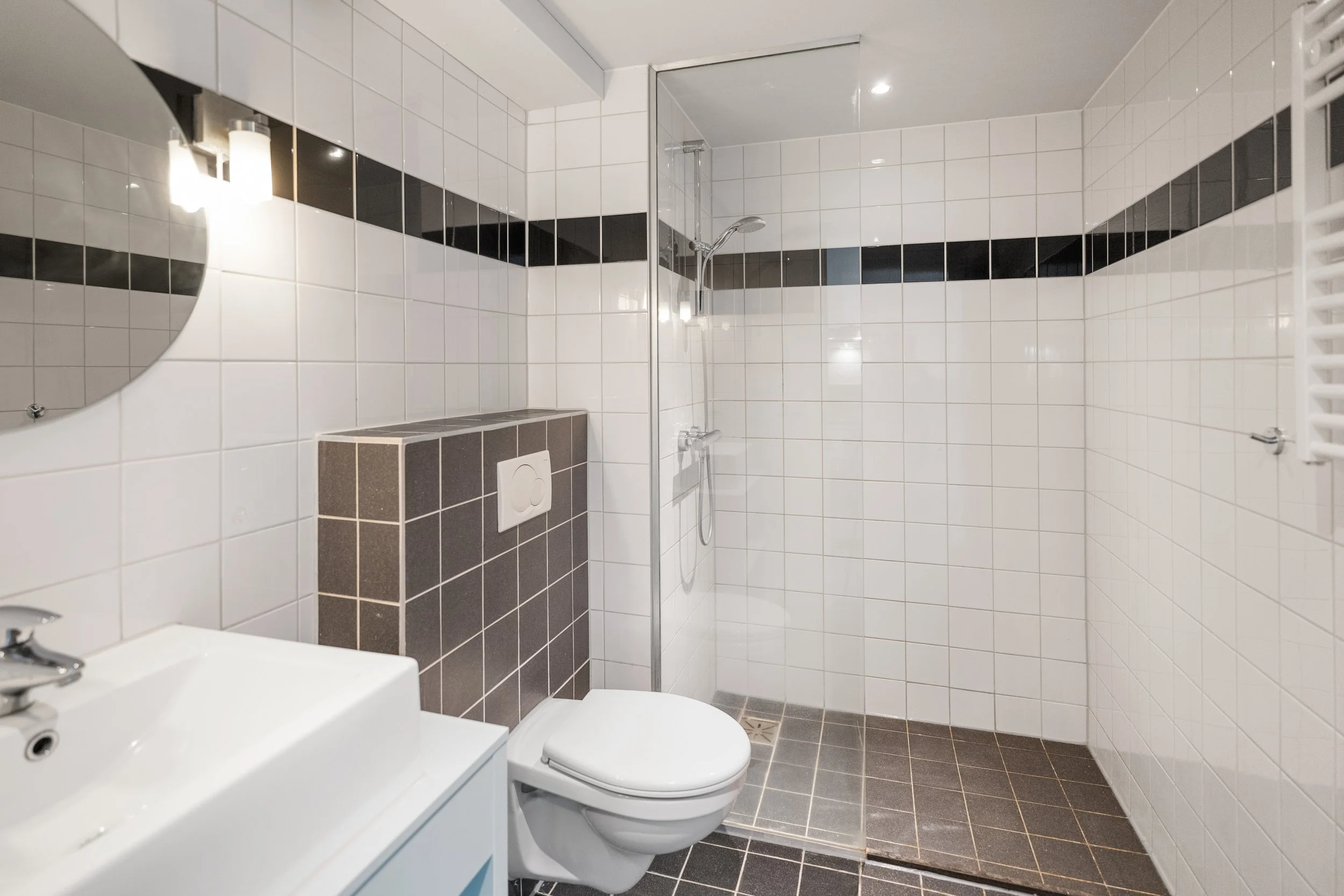Modern bathroom with white tiled walls, black accent tiles, a wall-mounted toilet, a shower area with a glass partition, a small white sink, and a round mirror.