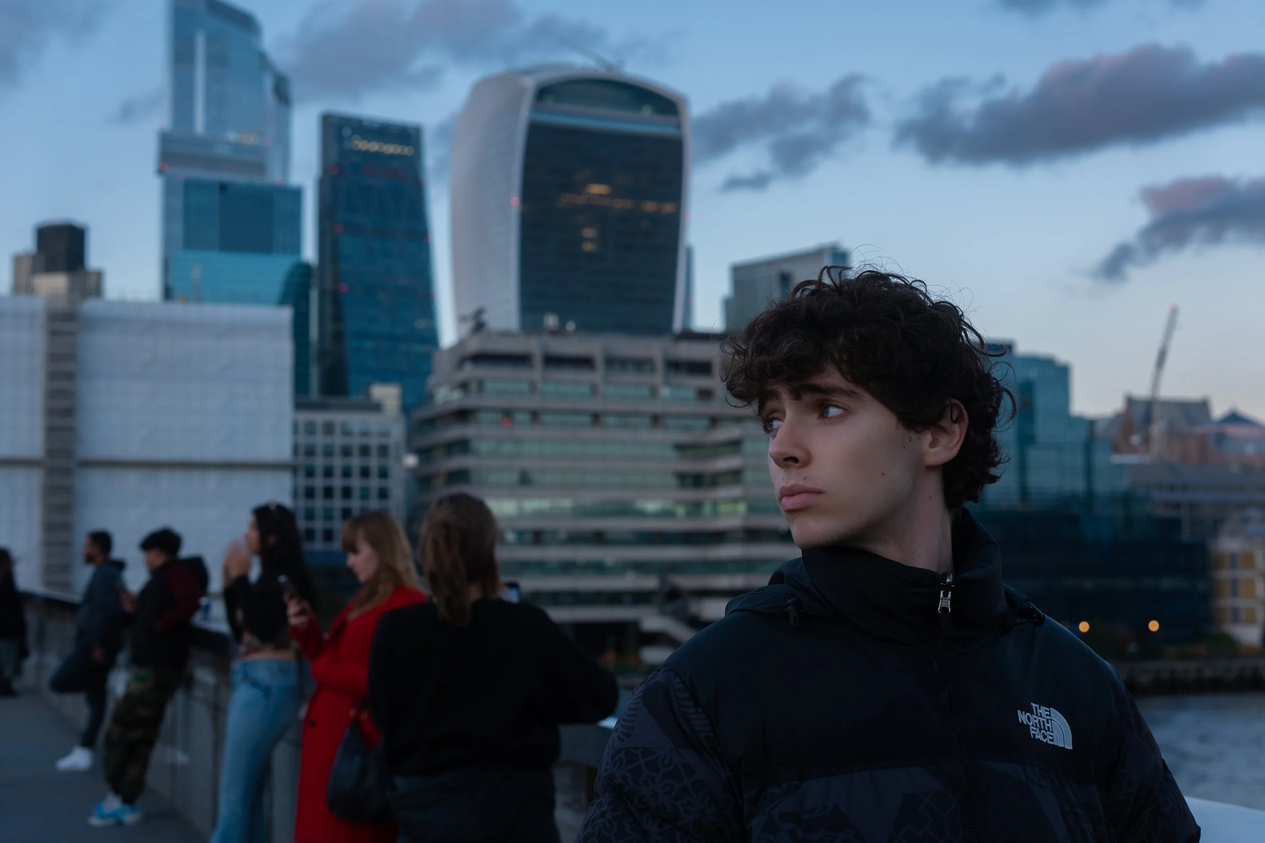 A young man with curly hair wearing a black North Face jacket stands on a bridge in London, with skyscrapers and a river in the background at dusk.