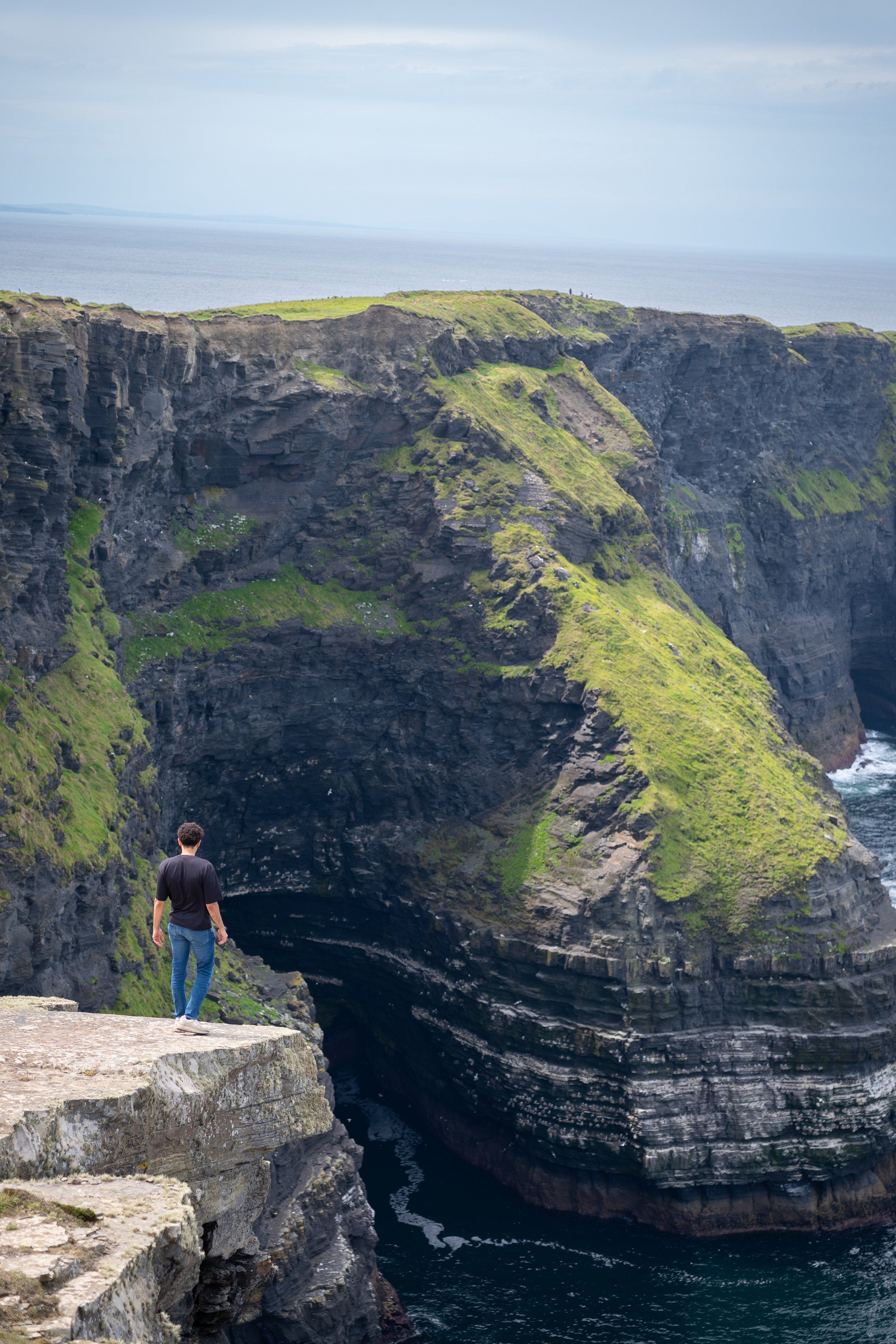 A person standing on a rocky cliff overlooking a large sea cave with green moss-covered cliffs and the ocean in the background.