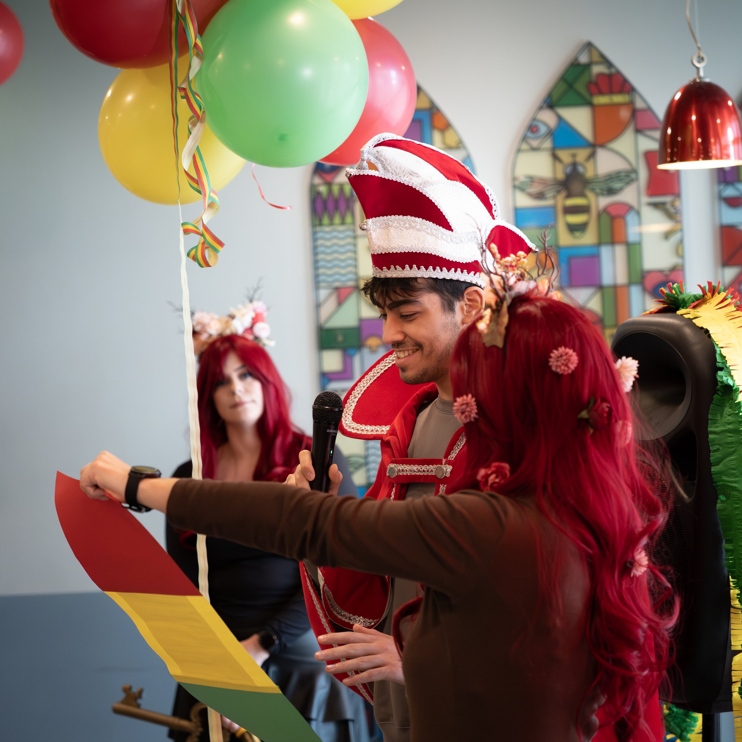 People celebrating a festive event indoors, with balloons, colorful decorations, and a man wearing a clown or festive hat.