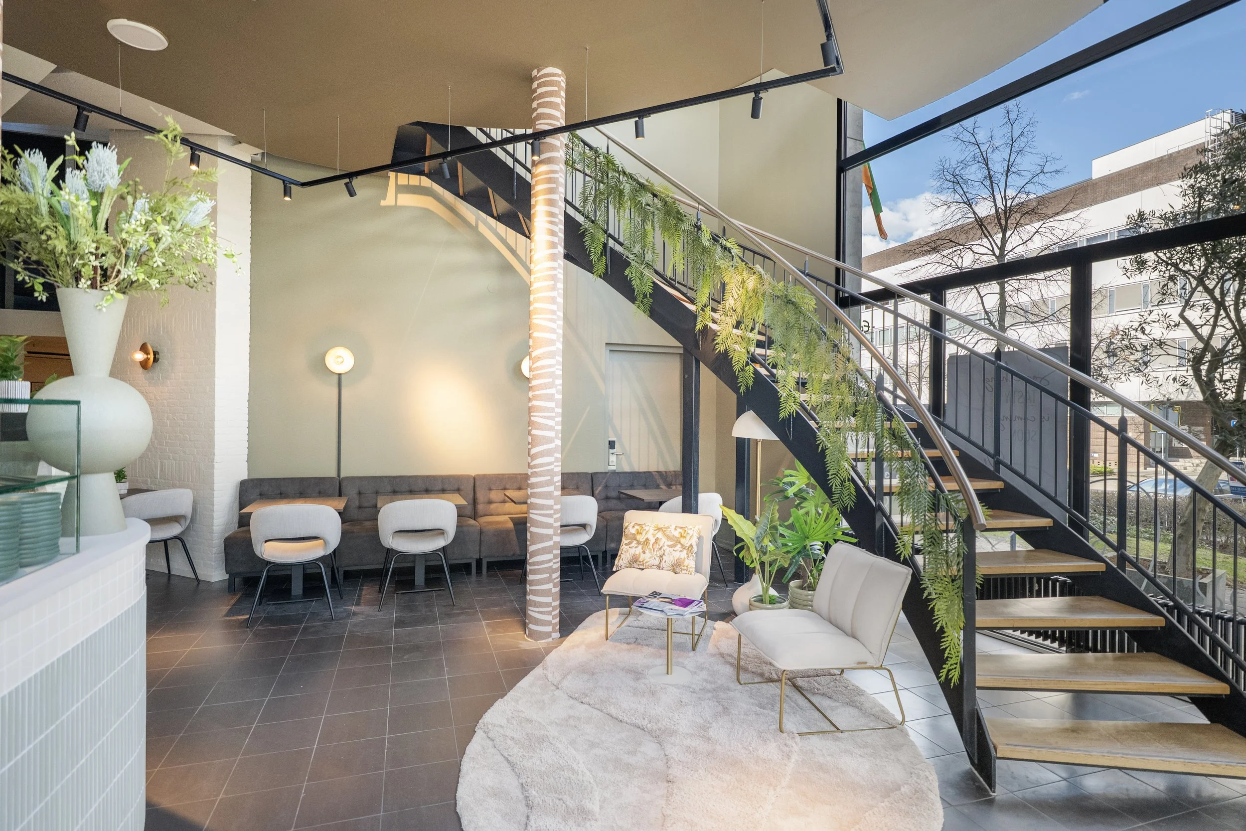 Modern interior of a cafe or lounge with a staircase decorated with hanging green plants, white chairs, a gray sofa, and large windows showing a city street outside.