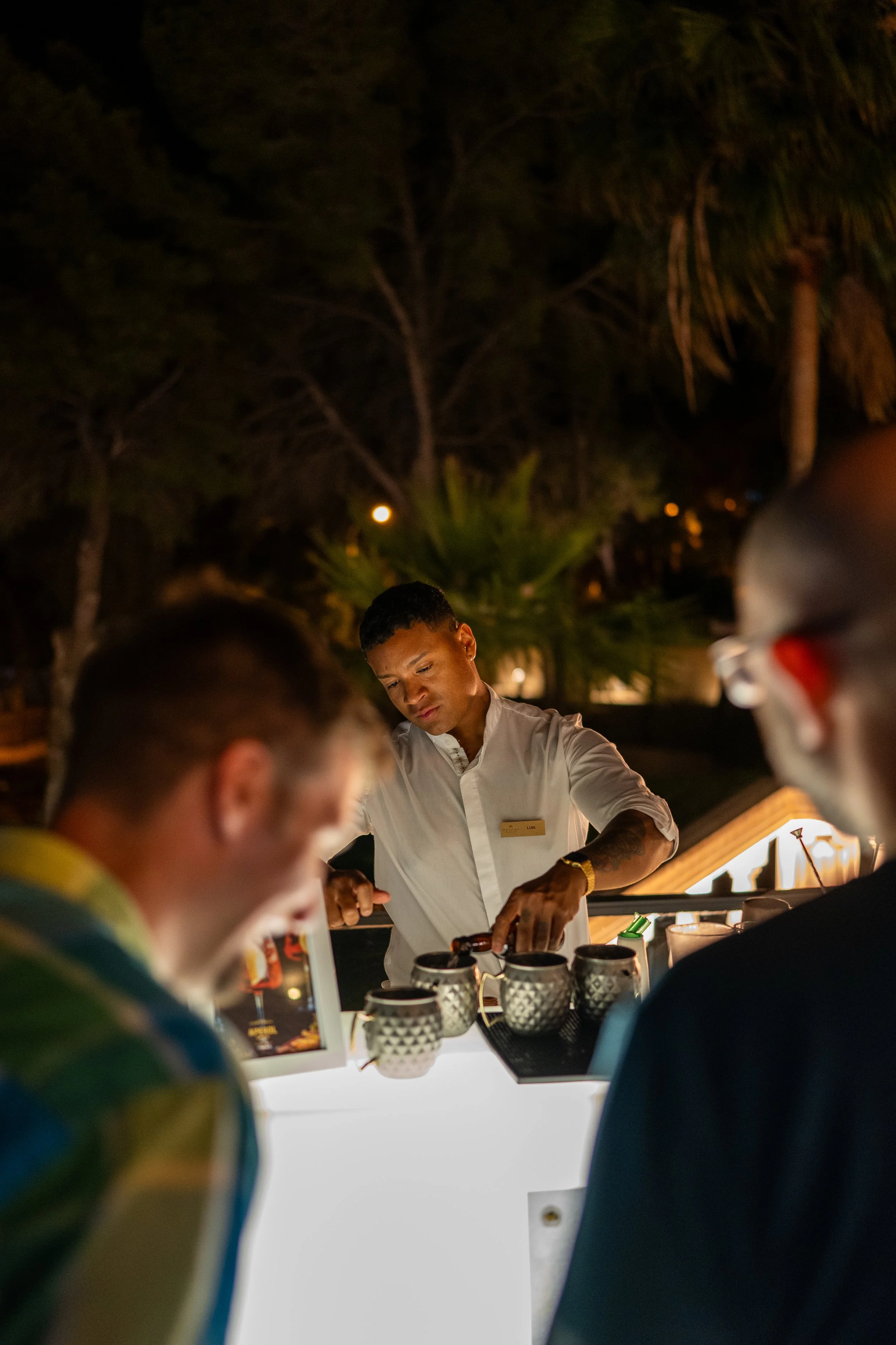 A bartender preparing drinks at an outdoor bar at night, with two customers in the foreground and palm trees and dark sky in the background.