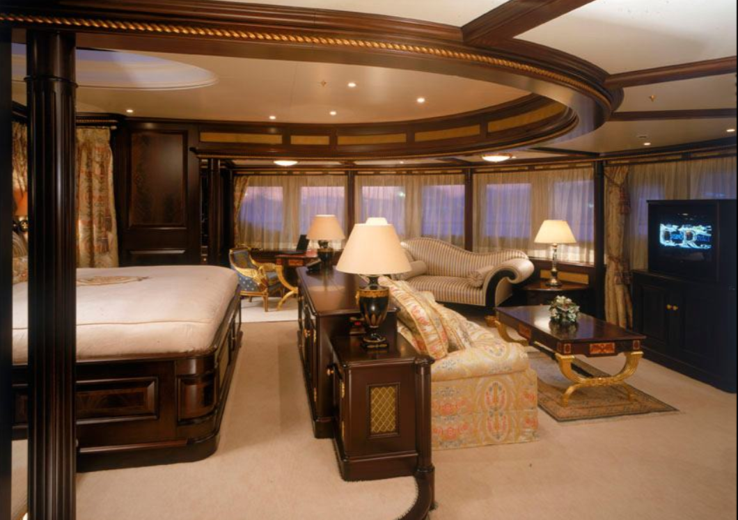 Luxurious hotel suite with a large bed, sitting area with striped sofa, side tables with lamps, a TV, and a panoramic window with curtains.
