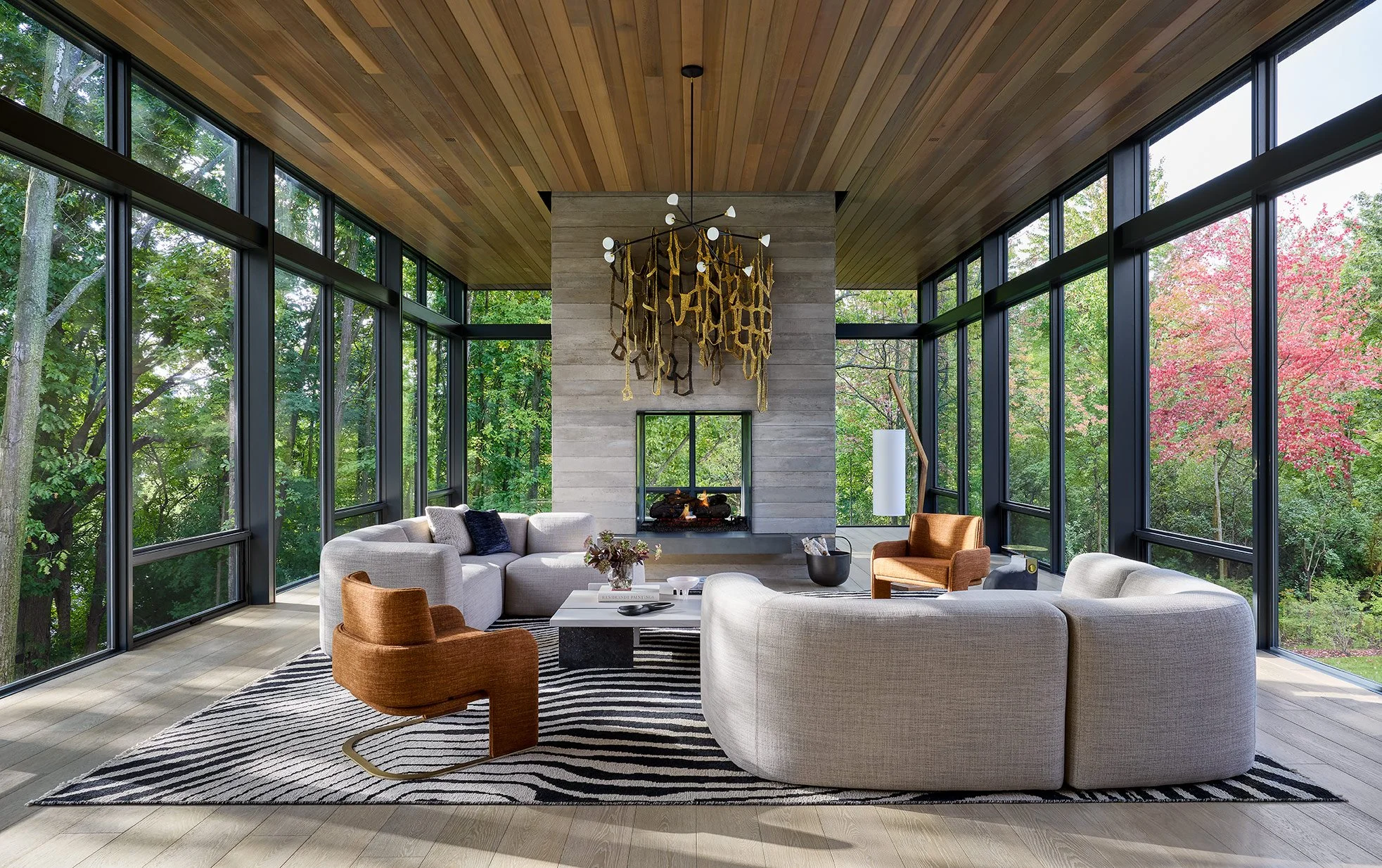 Modern living room with large glass windows showing green trees outside, neutral-colored curved sofas, a rust-colored armchair, a patterned rug, a fireplace, and a unique chandelier hanging from a wooden ceiling.