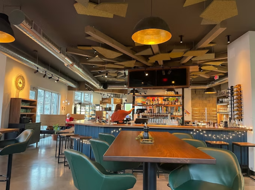 Interior of a modern coffee shop or cafe with a bar counter, green chairs, decorative ceiling panels, and large windows letting in natural light.