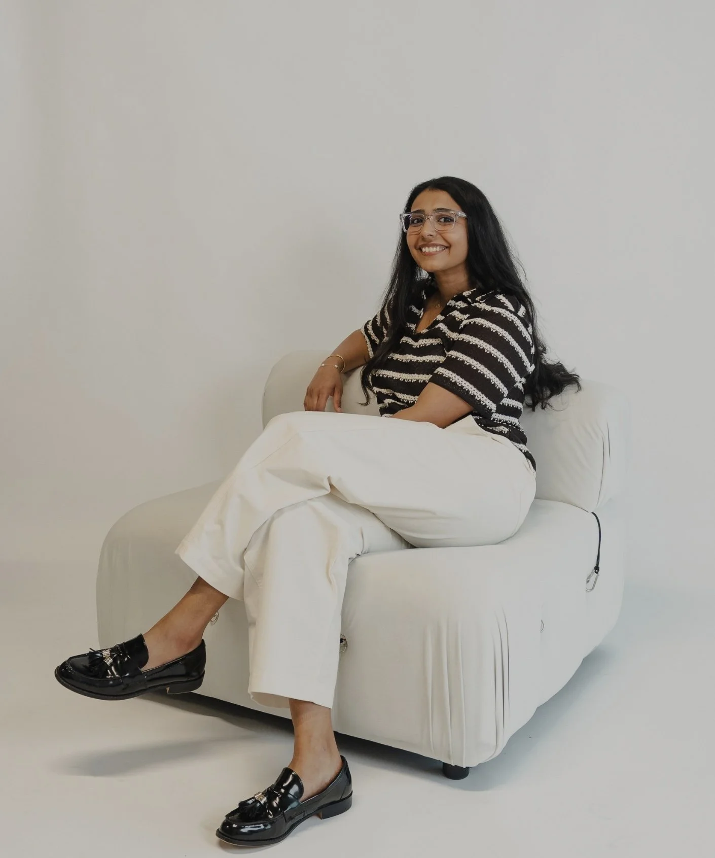 A woman with long black hair, wearing a gray sweater over a white top and black pants, sitting on a beige sofa in a room with a white wall and wooden floor, smiling at the camera.