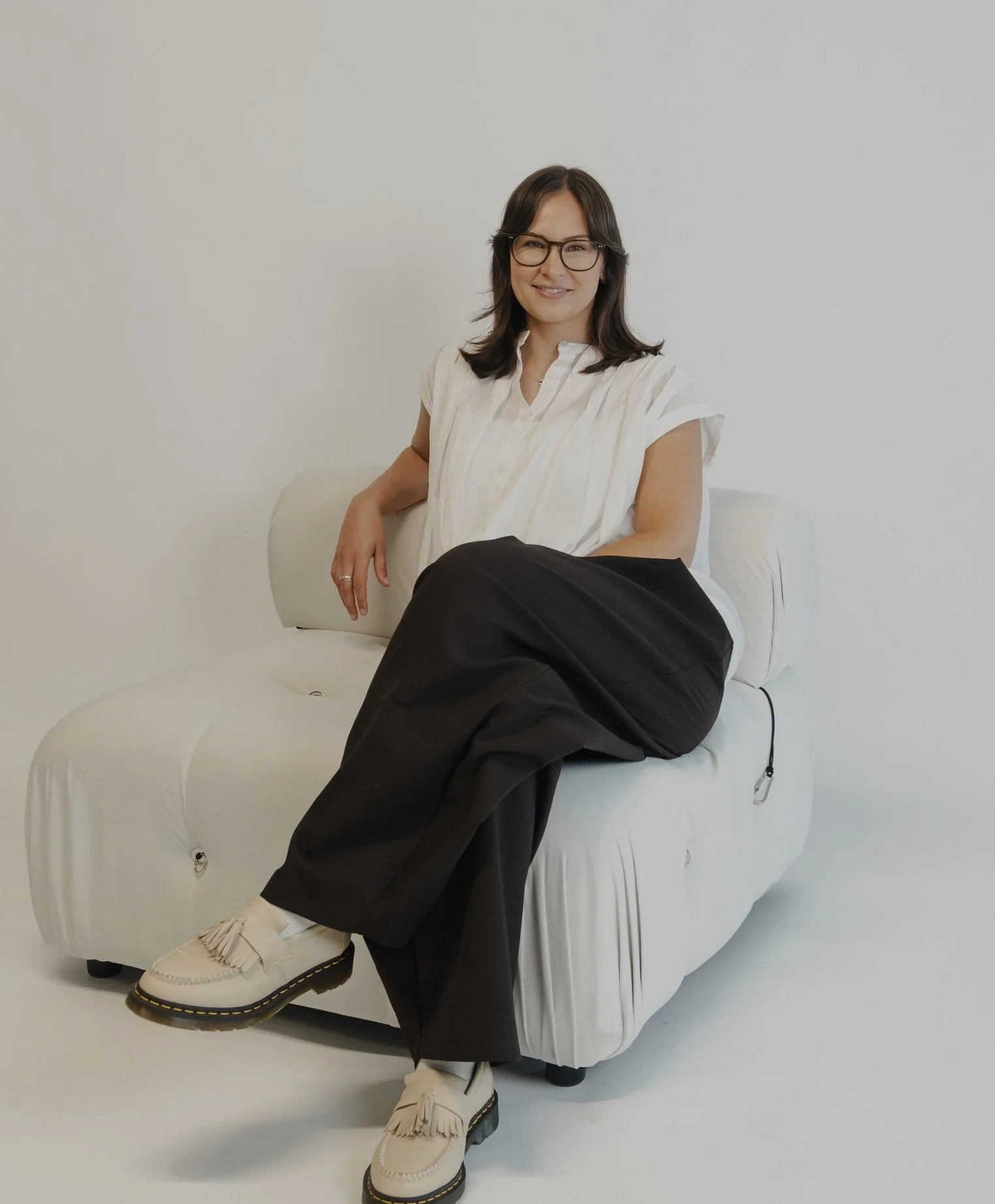 A woman with long dark hair, glasses, and a white striped shirt sits on a light-colored couch in a minimalist room, smiling gently.