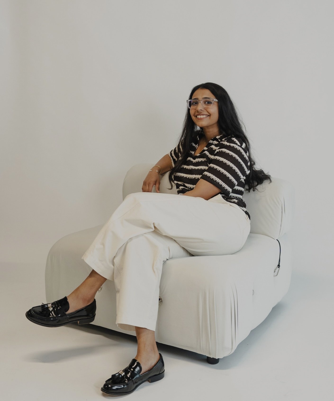 A woman with long dark hair sitting on a beige sofa, smiling at the camera, in a room with a white wall and wooden floor, wearing a gray cardigan and black pants.