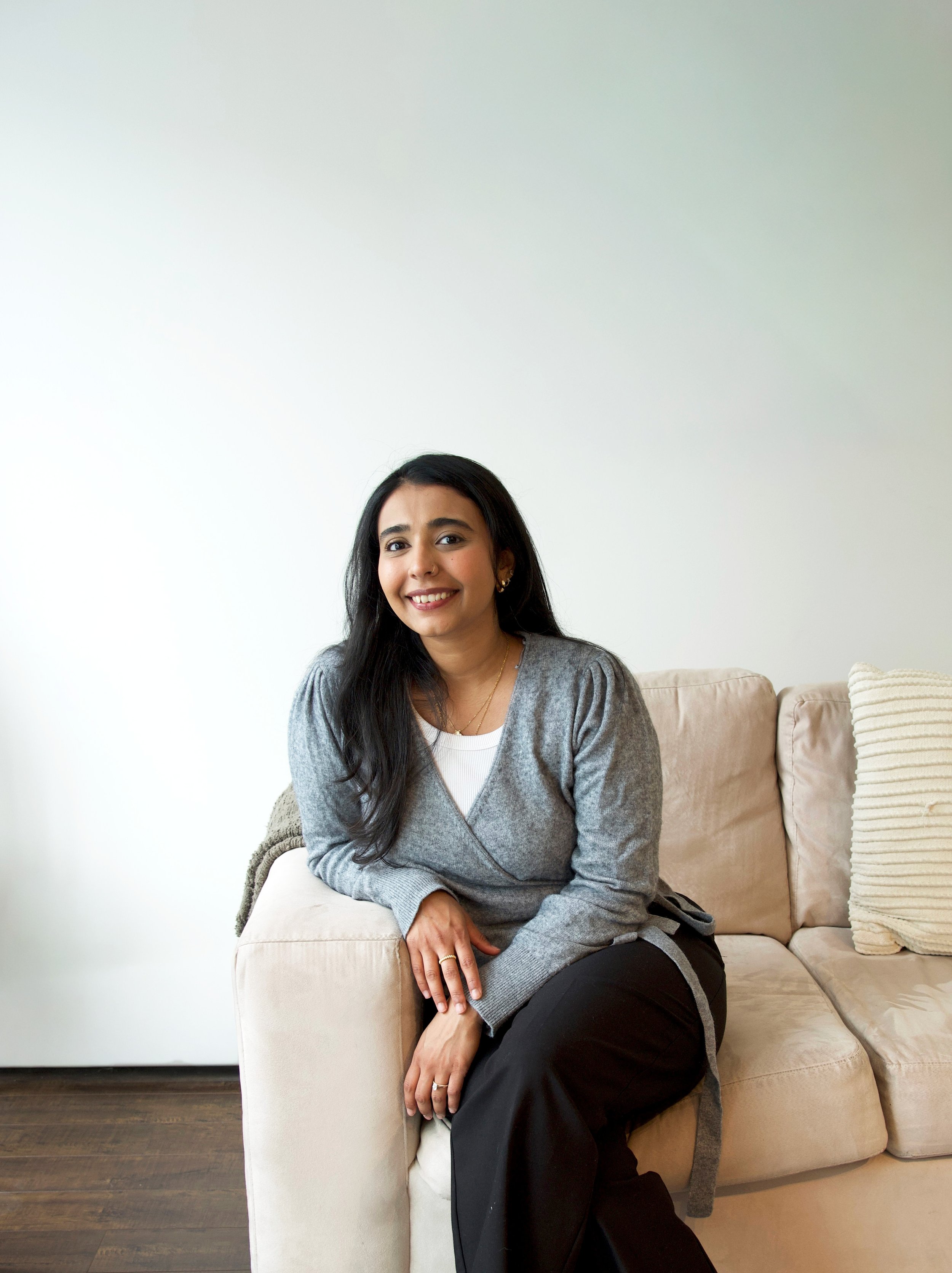 A woman with long black hair, wearing a gray sweater over a white top and black pants, sitting on a beige sofa in a room with a white wall and wooden floor, smiling at the camera.