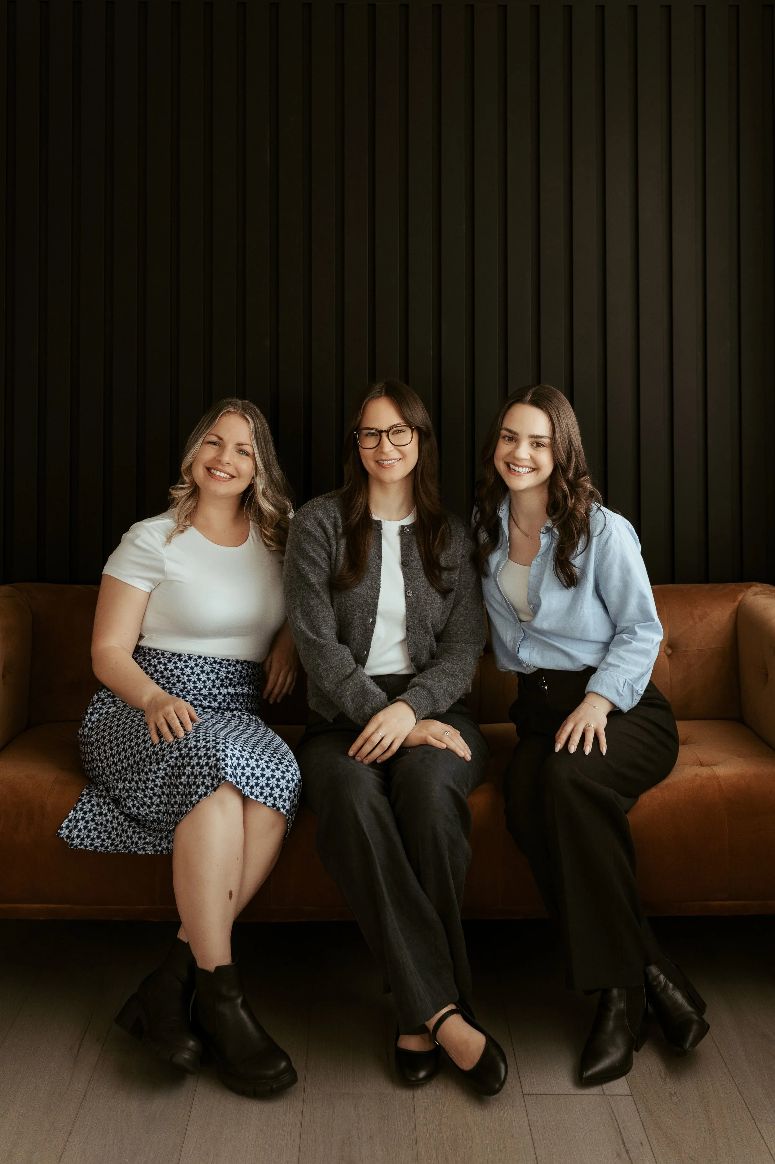 Three women sitting on a brown leather sofa against a black paneled wall, smiling at the camera.