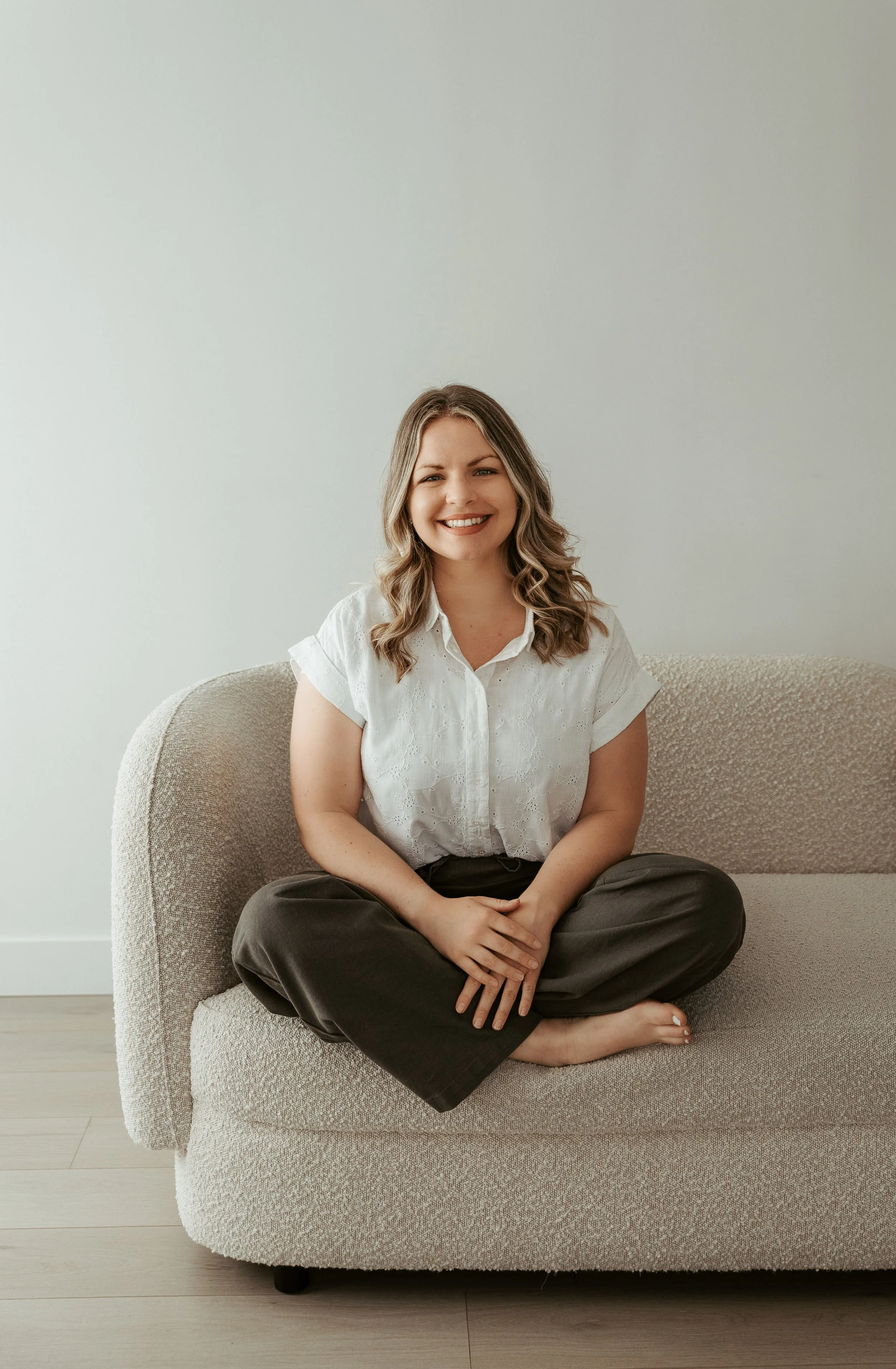 A woman with wavy brown hair, wearing a white short-sleeved blouse and black pants, sitting cross-legged on a beige sofa, smiling at the camera in a minimalistic room with a plain white wall behind her.