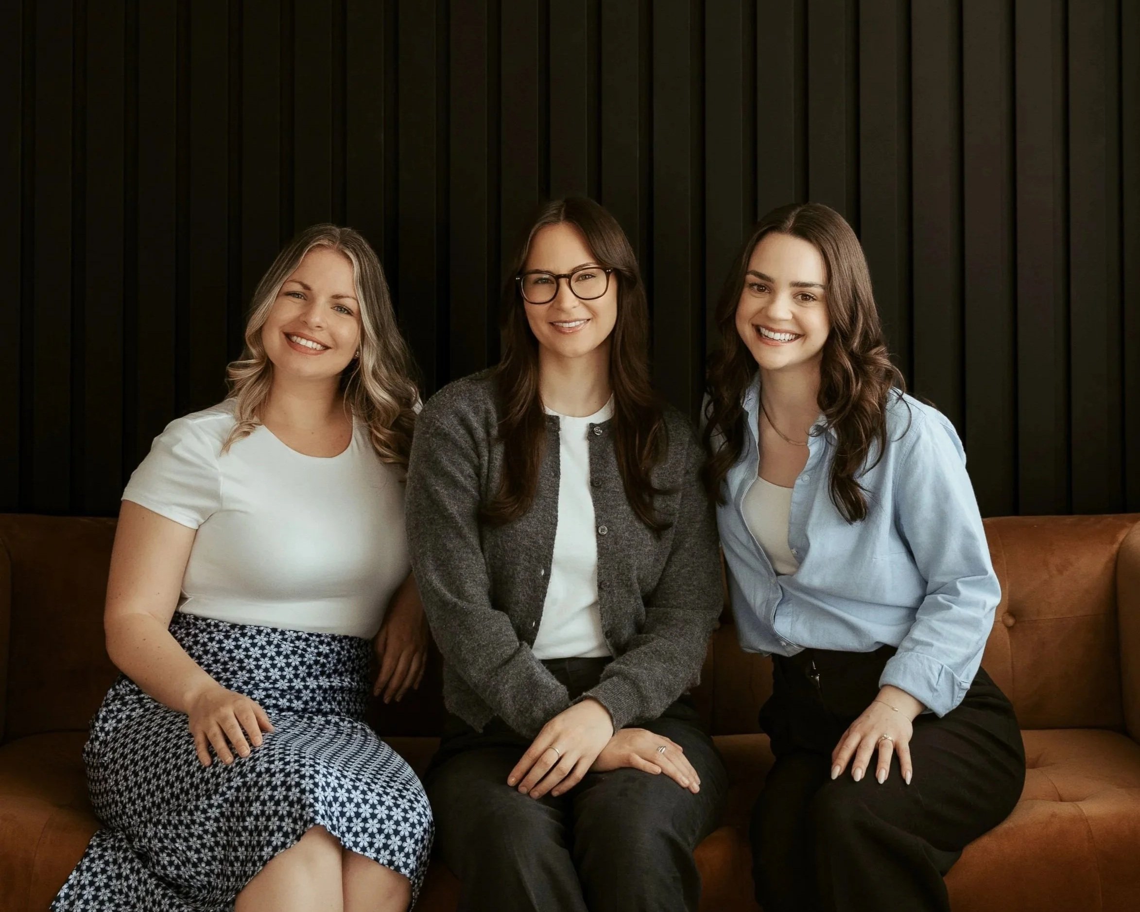 Three women sitting close together on a brown leather couch, smiling at the camera, with a black wooden wall behind them.